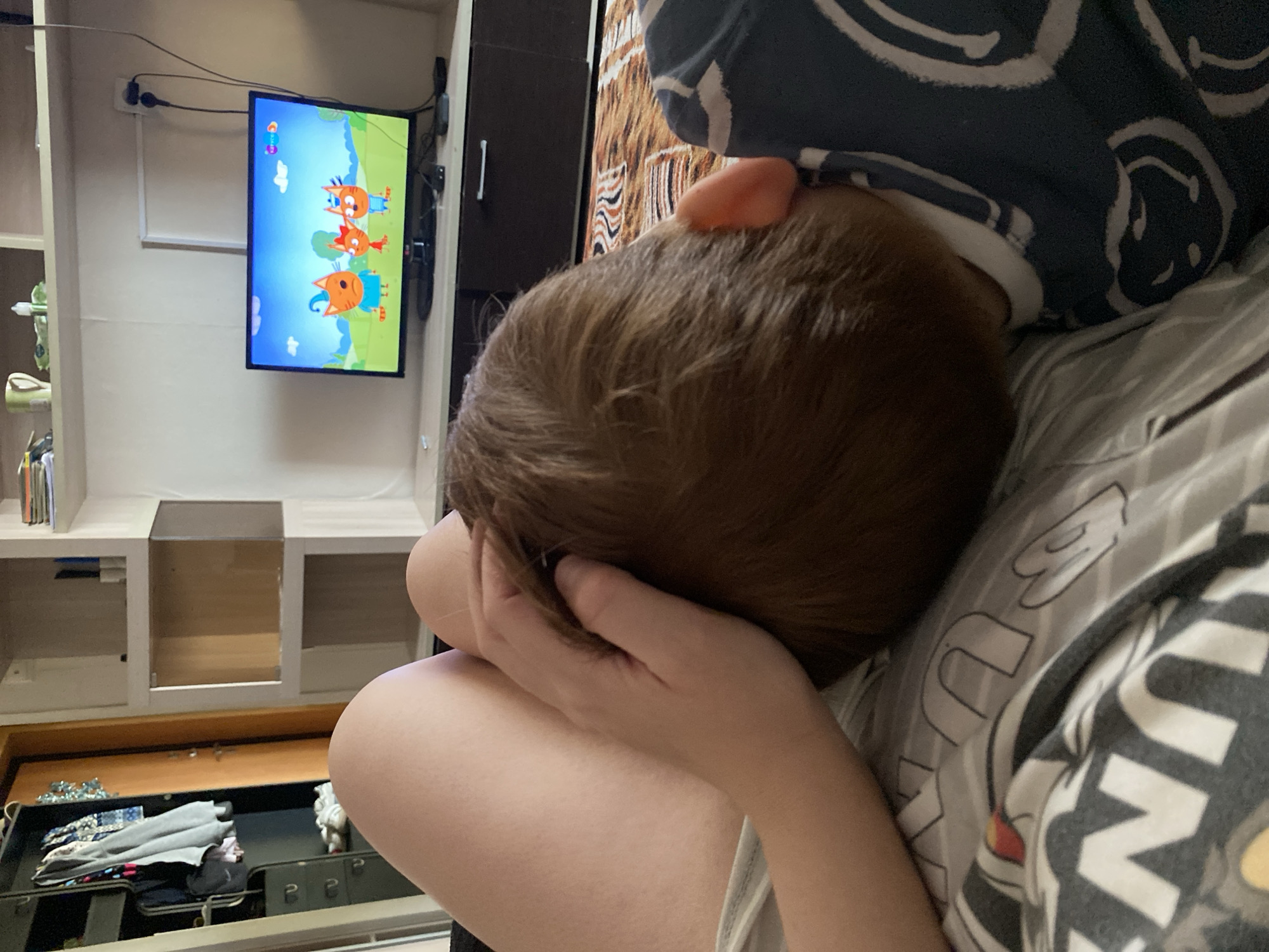 A child is lying down with their head resting on their arm. A television is mounted on the wall, displaying an animated show. A partially open dresser drawer contains clothes. The scene is located in an indoor environment, likely a home, in Chaikovsky, Russia.