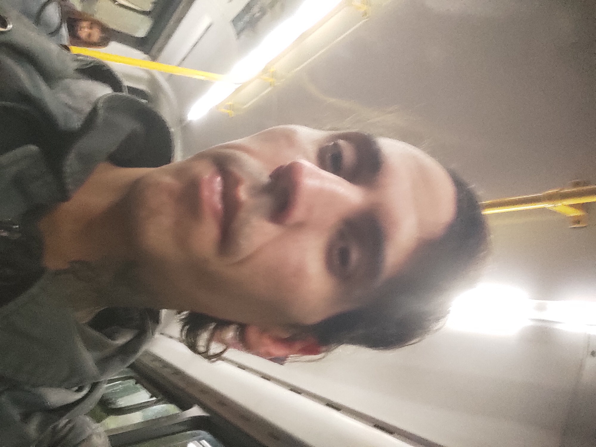 A man with dark hair and a gray hoodie is taking a selfie on a bus. Another person is visible behind him on the left. The bus interior is illuminated by overhead fluorescent lights. Based on the location given in the prompt, the bus is assumed to be in Gerli, Argentina.