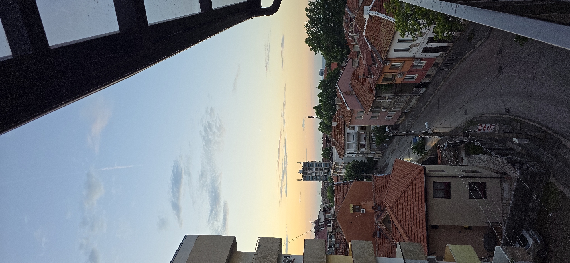 This image captures an urban landscape in Vratsa, Bulgaria, at either dawn or dusk, characterized by a gradient sky transitioning from light blue at the zenith to warm yellow and orange hues near the horizon.
In the upper portion of the frame, the sky exhibits scattered, sparse clouds and a prominent, linear contrail extending vertically. A small airplane is visible in the mid-right sky. A dark, possibly metal, architectural structure with square cutouts occupies the upper right corner, partially obscuring the sky, with a visible gutter running along its lower edge. On the far left, a section of a multi-story building with beige-colored textured facades and balconies is visible.
The midground and foreground depict a densely built residential area. Numerous buildings feature terracotta tiled roofs, varying in shape and pitch. The buildings themselves are a mix of styles, with some appearing older, constructed from materials like plaster and painted in muted tones such as pink, beige, white, and a darker reddish-brown. Many buildings have multiple windows and some are equipped with external air conditioning units. A winding, asphalt street, appearing damp or wet, traverses the scene from the lower center towards the mid-left. Utility poles with multiple overhead power lines are prevalent along the street. On the left side of the street, a light-colored sedan is parked near a two-story house with a reddish-brown tiled roof and dark-framed windows. Adjacent to this house, a red and white rectangular sign on a utility pole displays the word "OUTLET" in English and Bulgarian text below, indicating "SALES OF EDIBLE FATS AND OILS ELECTRICAL APPLIANCES DRUGSTORE".
In the background, a taller, multi-story building, appearing light blue or grey, stands centrally with a distinctive top structure that may include antennas or other technical equipment. To its right, a reddish communication tower is visible above the tree line. The distant horizon reveals a hazy, blue-hued landscape, likely mountains or distant plains, consistent with Vratsa's geographical setting. Dense green trees are interspersed throughout the urban fabric, particularly along the street and in the background.
No individuals or active human interactions are depicted in the scene. The overall impression is one of a quiet residential area at a transitional time of day.