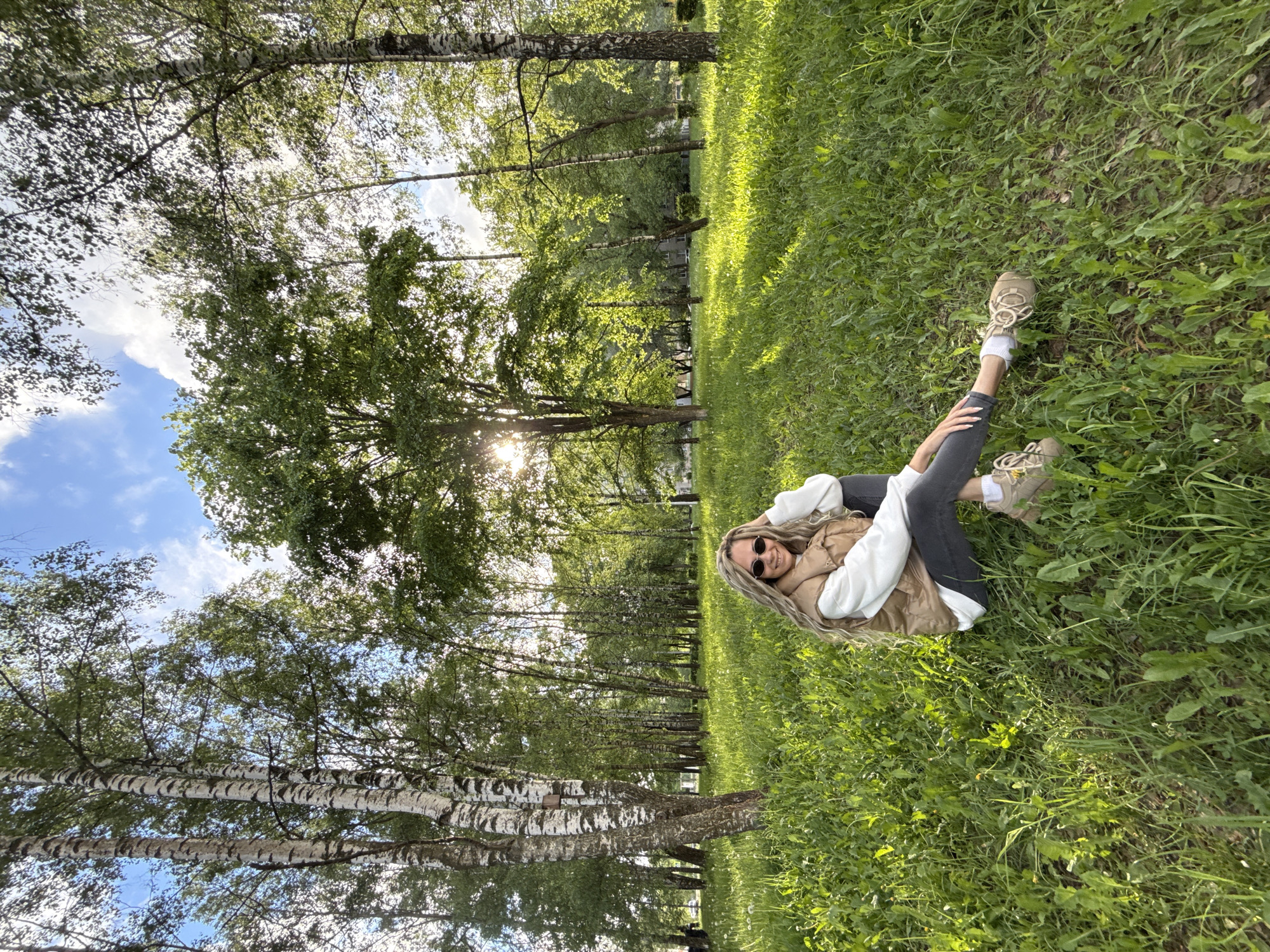 A woman is lying on her back in a grassy area within a park or wooded area. The location is assumed to be Rusanovo, Russia. She is wearing a white top, a brown vest, dark-colored pants, white socks, and tan sneakers. She is wearing sunglasses and has blonde hair. The area around her is covered in green grass. Trees surround the grassy area, with light filtering through the foliage. The sky is visible through the trees, showing some blue and white clouds.