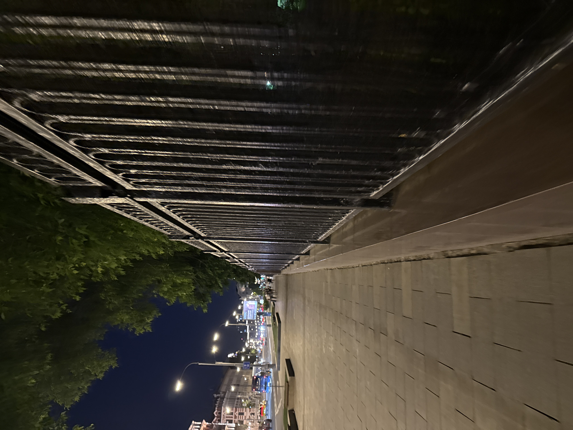 This nighttime image depicts a wide urban thoroughfare in central Moscow, Russia, specifically along the perimeter of Alexander Garden, adjacent to the Kremlin.
In the immediate foreground and extending into the mid-ground on the right, a dark, ornate metal fence with vertical bars and decorative arched tops runs parallel to the perspective. The fence's surface reflects artificial light, creating vertical highlights. Below the fence, a dark plinth separates it from a paved sidewalk. The sidewalk, composed of light-colored rectangular tiles, occupies the left-center of the foreground and extends into the distance.
To the left of the sidewalk, a multi-lane road is visible, illuminated by numerous streetlights positioned along its length. Vehicle headlights and taillights indicate active traffic. A blue public bus and several passenger cars are present on the road in the mid-ground. Further down the street, on the left, a large, brightly illuminated billboard is prominent.
Across the road, a line of multi-story buildings with classical architectural elements, including ornate facades and arched windows, are illuminated. In the distance on the left sidewalk, a small group of pedestrians can be observed walking. Overhead, in the upper left portion of the frame, dense green tree foliage partially obscures the dark night sky. The scene is lit predominantly by artificial streetlights and building illumination, casting a warm glow on the pavement and highlighting urban structures.