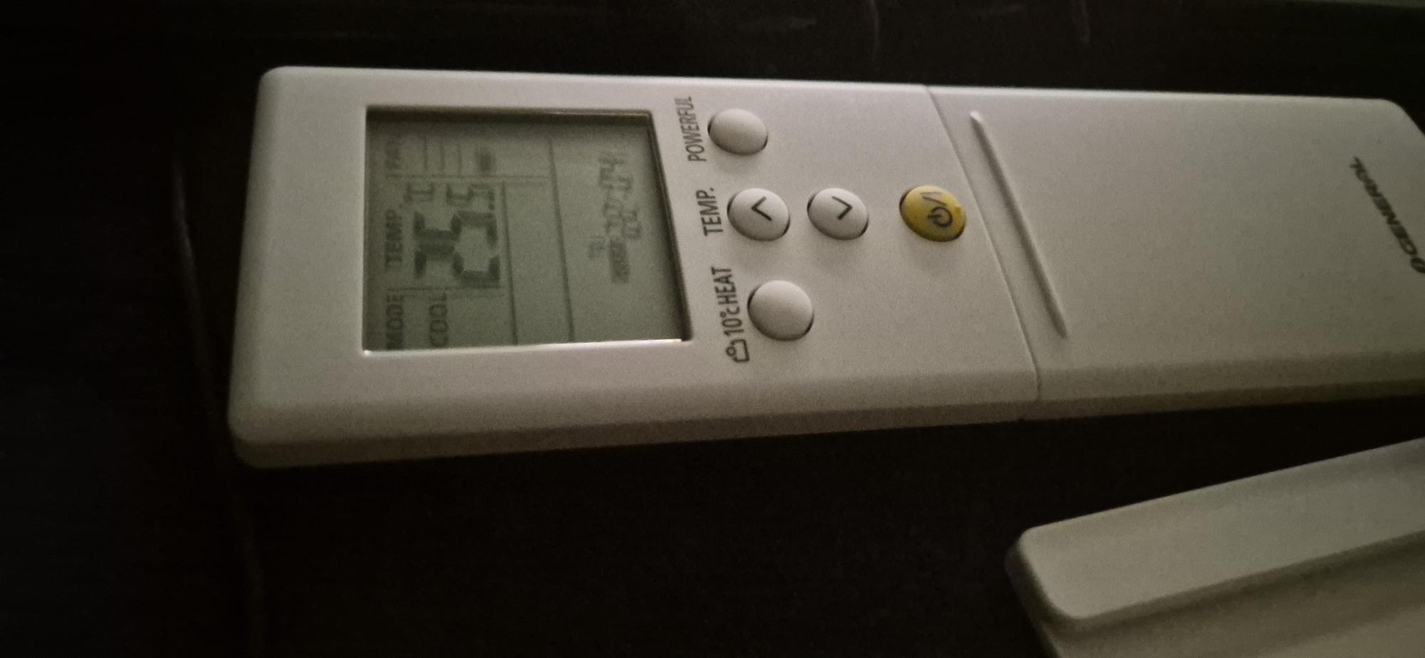 The image is a close-up, high-angle shot of a white remote control for an air conditioner. The remote is lying on a dark, textured surface. The remote control has a digital display showing "25.5" and other symbols. Buttons on the remote include "MODE," "COOL," "TEMP," "POWERFUL," and directional arrows, as well as a yellow power button with "ON/OFF" written on it. The brand name "GENERAL" is visible on the side of the remote. Another white object, possibly a cover or part of another device, is visible in the lower right corner of the image. The background is dark and out of focus.
