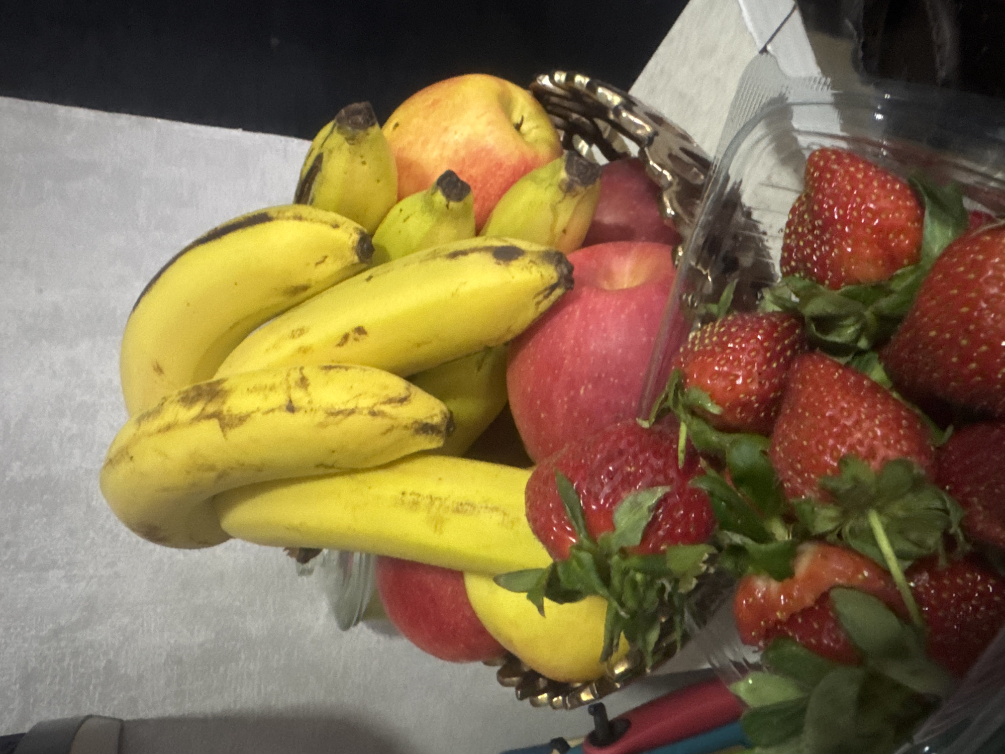 A bowl of fruit, including bananas, apples and strawberries, sits on a light gray surface. The bananas are yellow with some brown spots. The apples are red and yellow. The strawberries are in a plastic container. The location is unknown.
