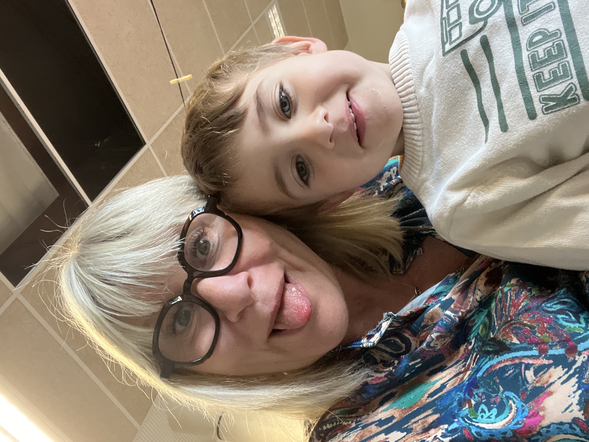 The image depicts a selfie taken from a low angle, featuring an adult female and a young child. The woman has short, blonde hair and wears black-framed glasses. She is sticking her tongue out. The child, with light brown hair and blue eyes, is smiling. He is wearing a white sweatshirt with a green design that includes the word "KEEP". The woman is wearing a colorful patterned shirt. The background is a tiled wall. There is no evidence of La Fouillouse, France in the image.