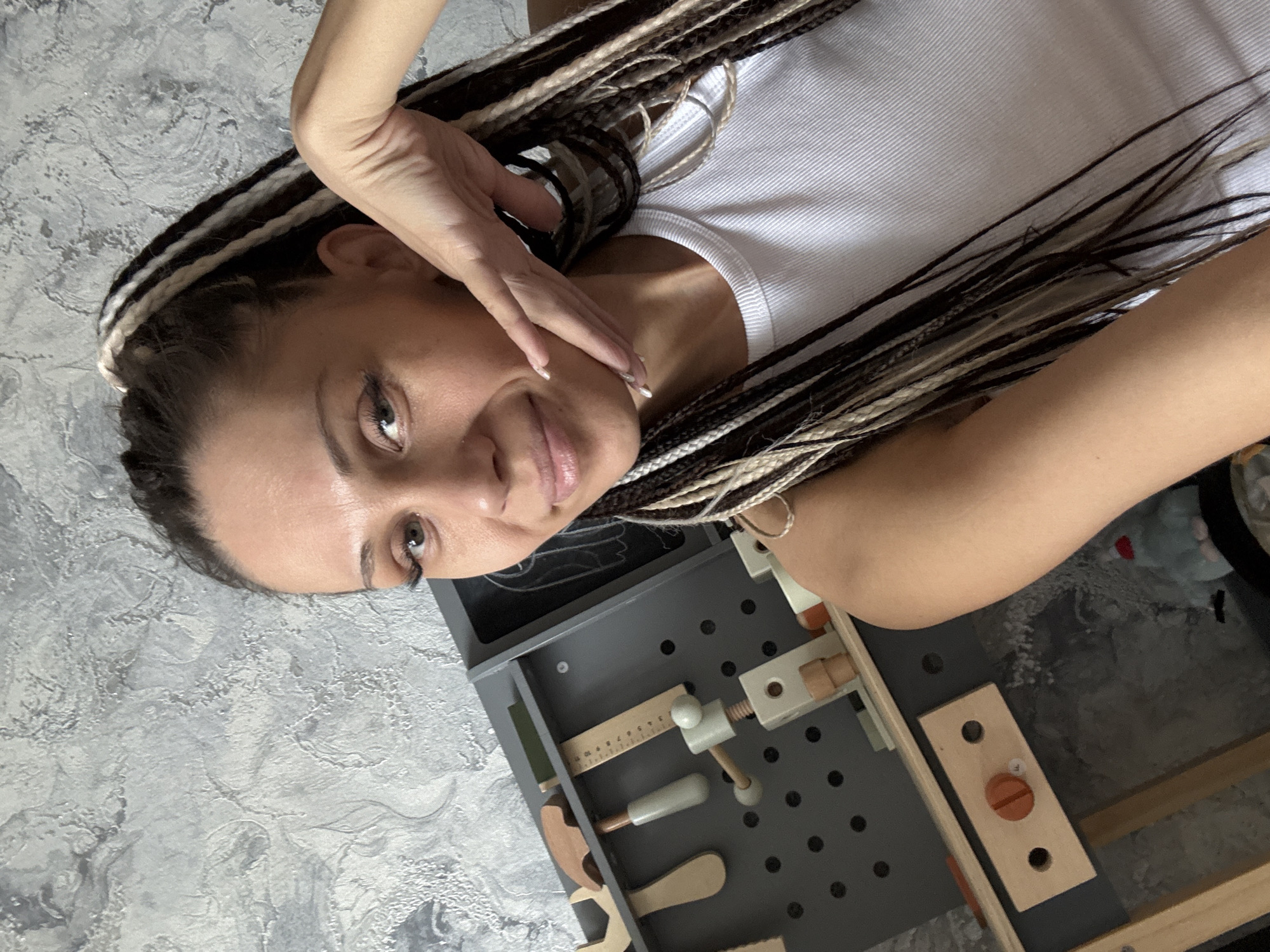 A woman with braided hair, a white tank top, and light skin is positioned at an angle, with her face close to the camera. She is touching her face with her hand. In front of her is a gray workbench with various wooden tools, including a ruler, a vise, and a wrench. The surface beneath her is a light gray with a textured, possibly marble-like pattern. The location appears to be indoors. It is not possible to confirm that the location is Rusanovo, Russia from the image.