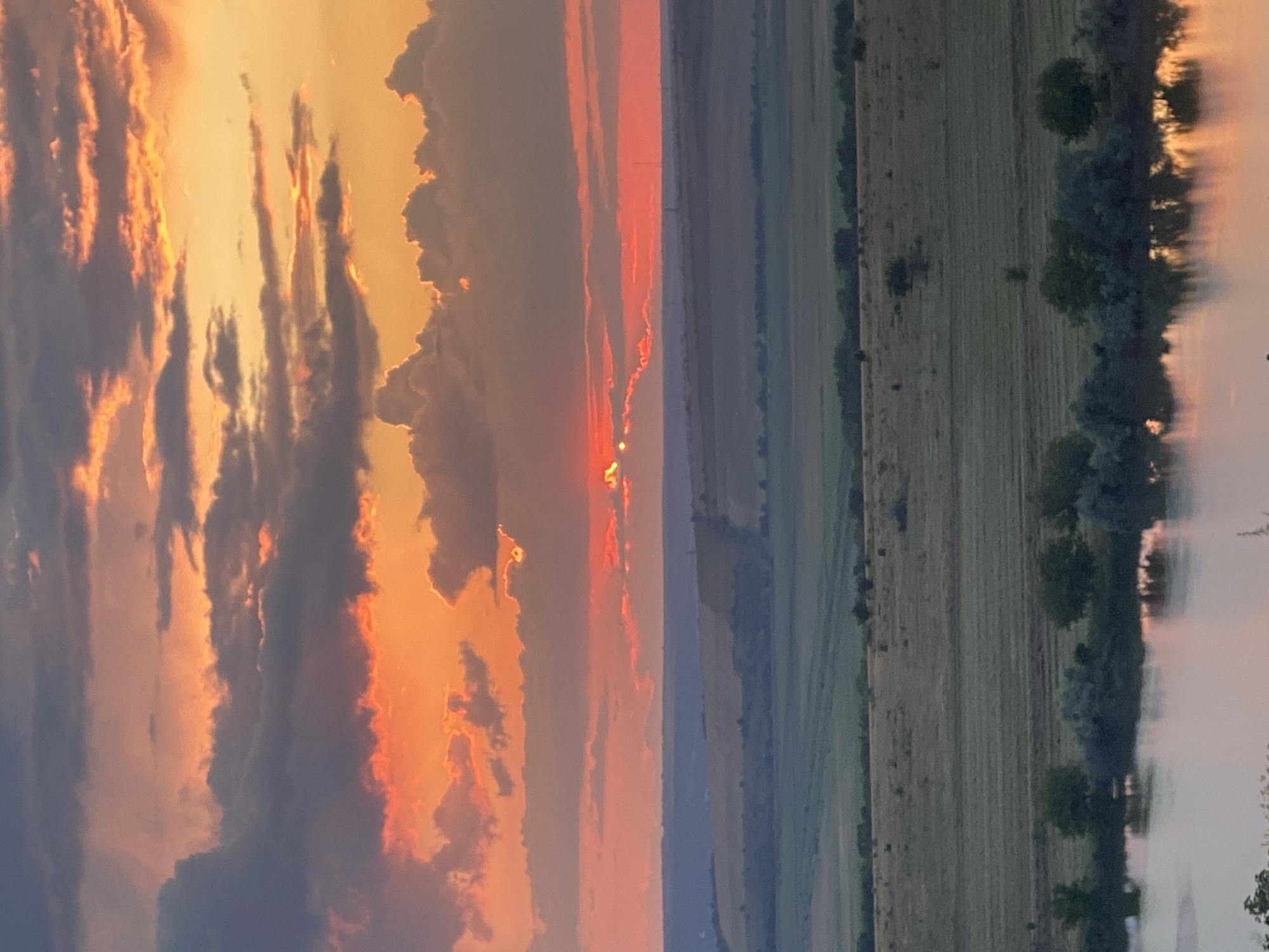 The image captures a dramatic sunset over a rural landscape near Cairaclia, Moldova. The sky is ablaze with vibrant hues of orange, red, and yellow, with dark gray clouds adding contrast. The sun, partially obscured by a thin layer of clouds, is visible as two bright points of light. Below the sky, rolling hills covered in fields of dry grass and green crops stretch towards the horizon. A body of water, likely a river or lake, occupies the foreground, reflecting the colors of the sunset. Trees and bushes line the water's edge, with scattered trees dotting the fields. The overall scene conveys a sense of natural beauty and tranquility at the end of the day.