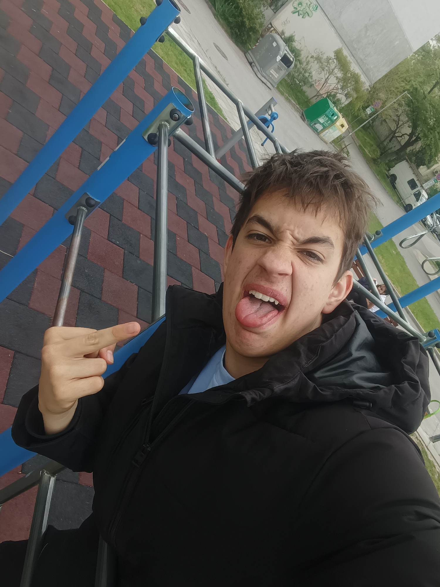 A young person is in a selfie-style photograph, making a face and extending their middle finger. They are wearing a black jacket over a blue shirt. The person appears to be standing or sitting on a piece of playground equipment constructed of blue bars and metal. The ground under the structure appears to be composed of alternating rows of red and gray brick. In the background, there is a street with a white van, several bins, and trees. The location may be in Varna, Bulgaria, but additional details are needed to confirm this.