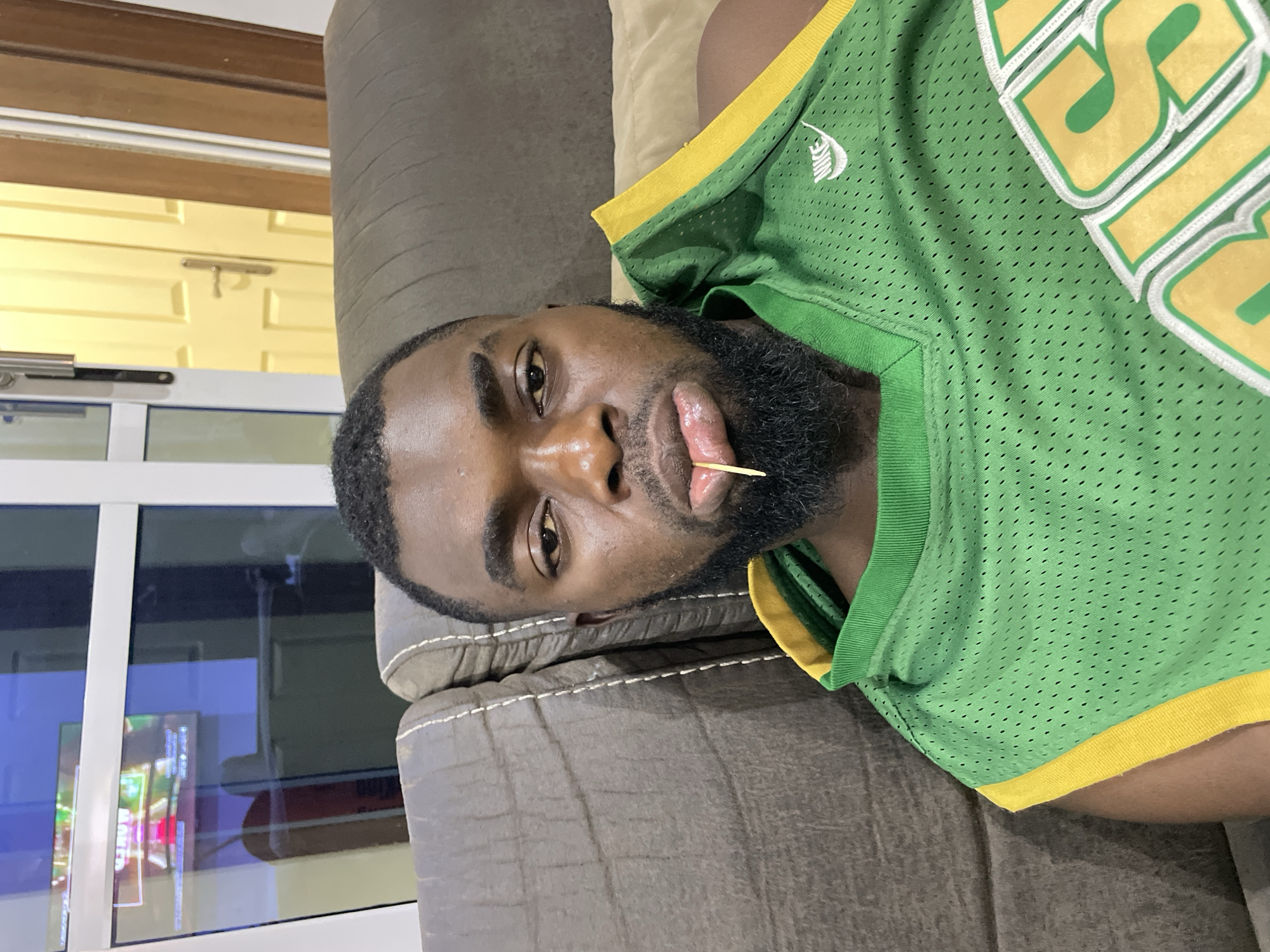 This media file features a dark-skinned man reclining on a sofa, captured indoors in Jos, Nigeria. He is seen from the chest up, with his head tilted to the right due to the image's orientation. The man has a full black beard and short, dark hair, and looks directly towards the camera with a neutral expression. A wooden toothpick is held between his lips.
He is wearing a bright green mesh basketball jersey with yellow trim around the neck and sleeves. A white "Nike" logo is clearly visible on the upper left chest area of the jersey. Large, yellow lettering, possibly an "S" or "JS", with a white outline, is partially visible on the front of the jersey.
The man is resting on a grey or brown upholstered sofa, which features visible white stitching. The background reveals elements of an indoor space: to the left, a light yellow door with visible paneling and a silver handle is present. Below this, a white-framed window reflects interior lights and what appears to be a colorful, fragmented image from a screen, possibly a television or monitor, suggesting the time of day might be evening or night. The scene conveys a sense of casual relaxation.
