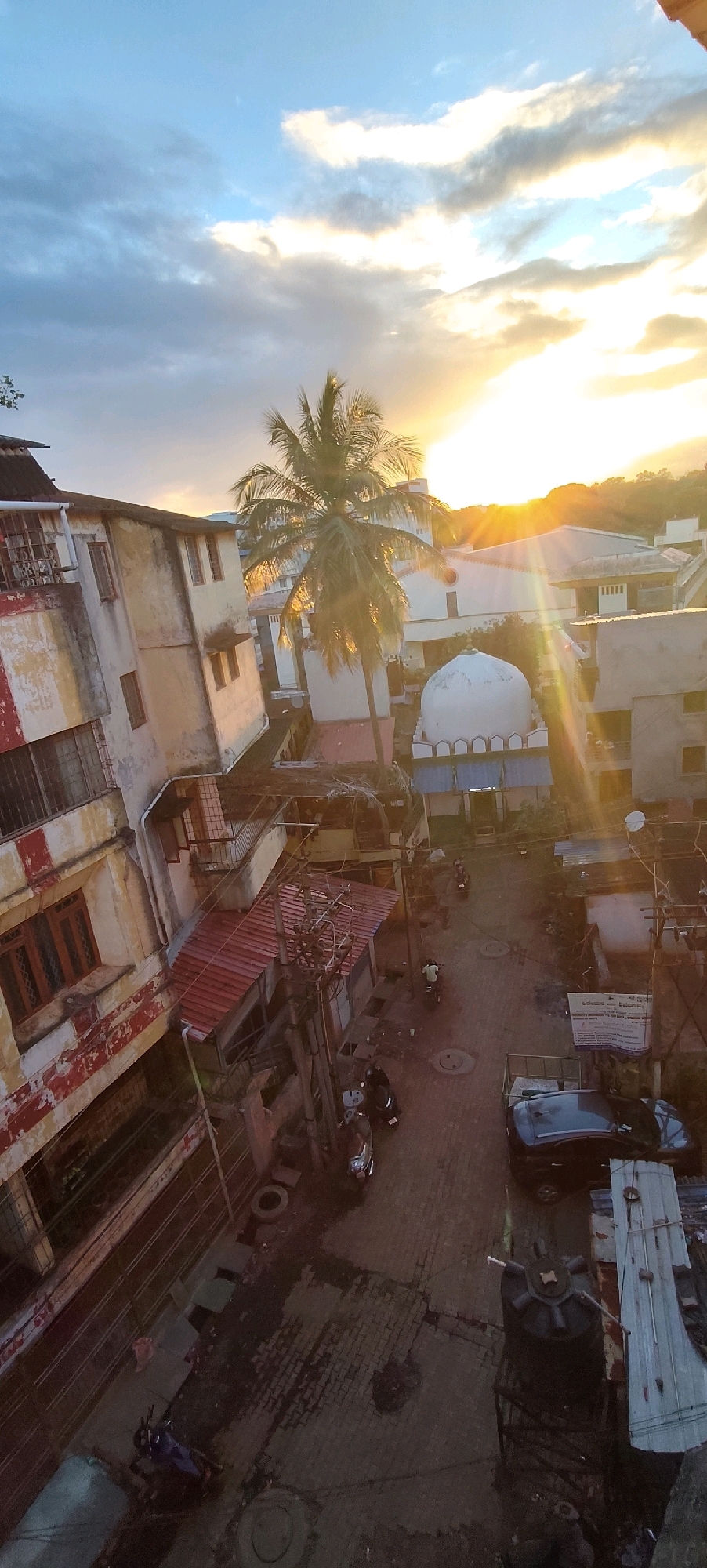 This aerial, slightly tilted, full-frame, high-angle shot captures a street scene in Belagavi, India, during sunset. The sky is a mixture of blue and grey clouds, with the setting sun casting a golden-orange glow and prominent sunbeams across the scene. In the foreground, a weathered, multi-story building with a red and white striped facade dominates the left side of the frame. Its windows are barred, and an attached balcony is visible. Adjacent to this building is another structure with a red corrugated metal roof.
A prominent palm tree stands in the middle ground, its fronds silhouetted against the bright sky. To the right of the palm tree is a white-domed building, likely a mosque, with arched windows and a blue-trimmed base. Beyond these structures, more buildings and a distant hillside are visible.
The street below is paved with brick and appears wet, suggesting recent rain. Several objects are present in the street: a black car parked on the right, a large black water tank on a raised metal platform, and a few motorcycles. One motorcycle is parked on the left, and another is ridden by a person wearing a white shirt and green pants in the middle of the street. A third person on a motorcycle, wearing a yellow shirt, is also visible further up the street. Electrical poles and wires crisscross the scene.
The overall impression is a lived-in urban environment at the end of the day, illuminated by the warm light of the setting sun. The image is sharp with good detail.