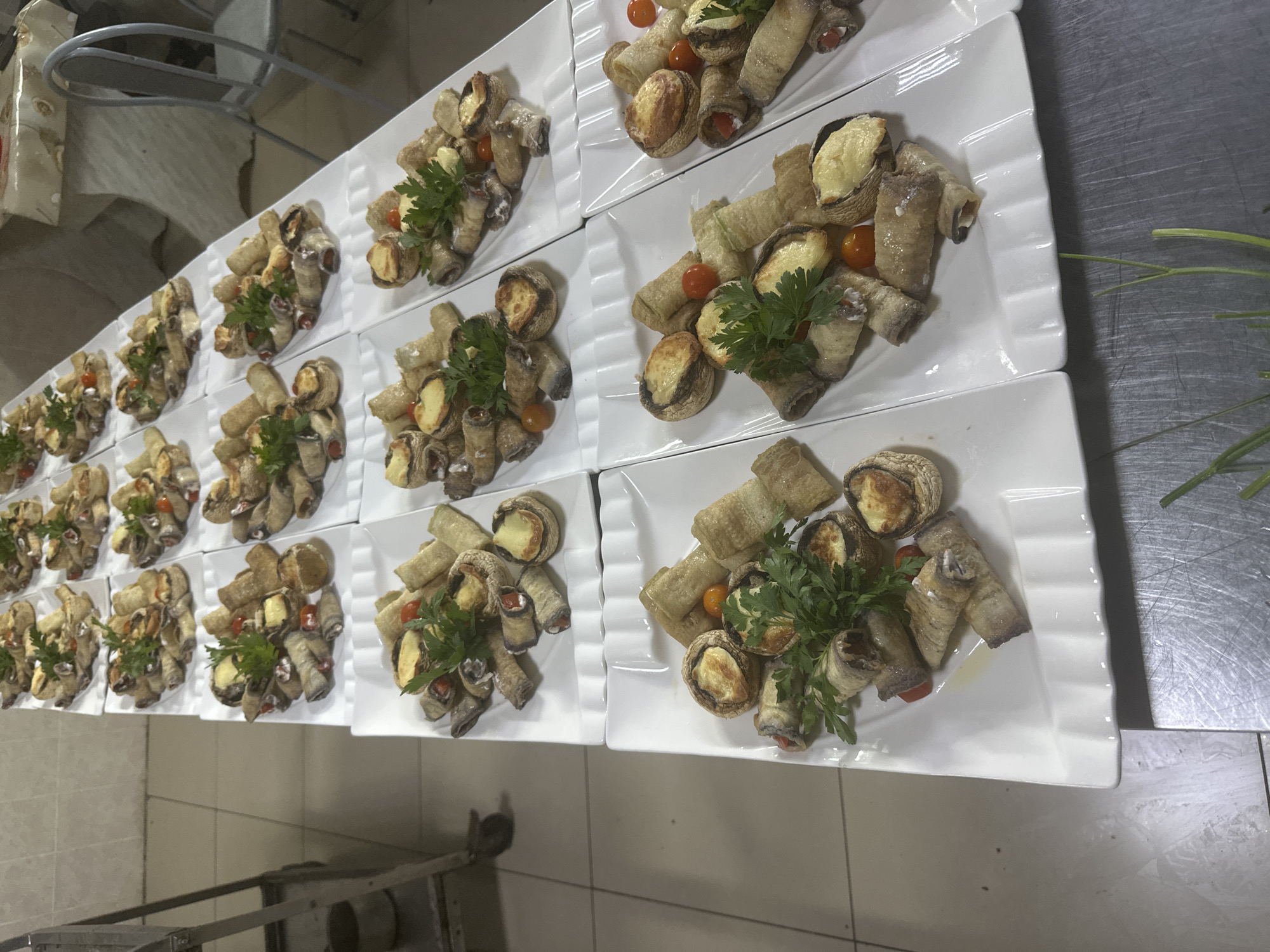 The image shows a catering setup with multiple white rectangular plates filled with appetizers. Each plate contains several pieces of what appear to be stuffed eggplant rolls, some stuffed mushrooms with a golden-brown topping, and small cherry tomatoes. Fresh parsley sprigs are used as a garnish on many of the plates. The plates are arranged in rows on a white surface, likely a table or counter, and some are partially obscured by others. In the background, a stainless steel kitchen cart is visible to the left, and chairs are present to the right. The overall impression is that of a prepared meal for an event. There are no people or specific landmarks visible that would indicate the city of Kostanay, Kazakhstan, or any other specific location. The environment appears to be an indoor kitchen or preparation area.