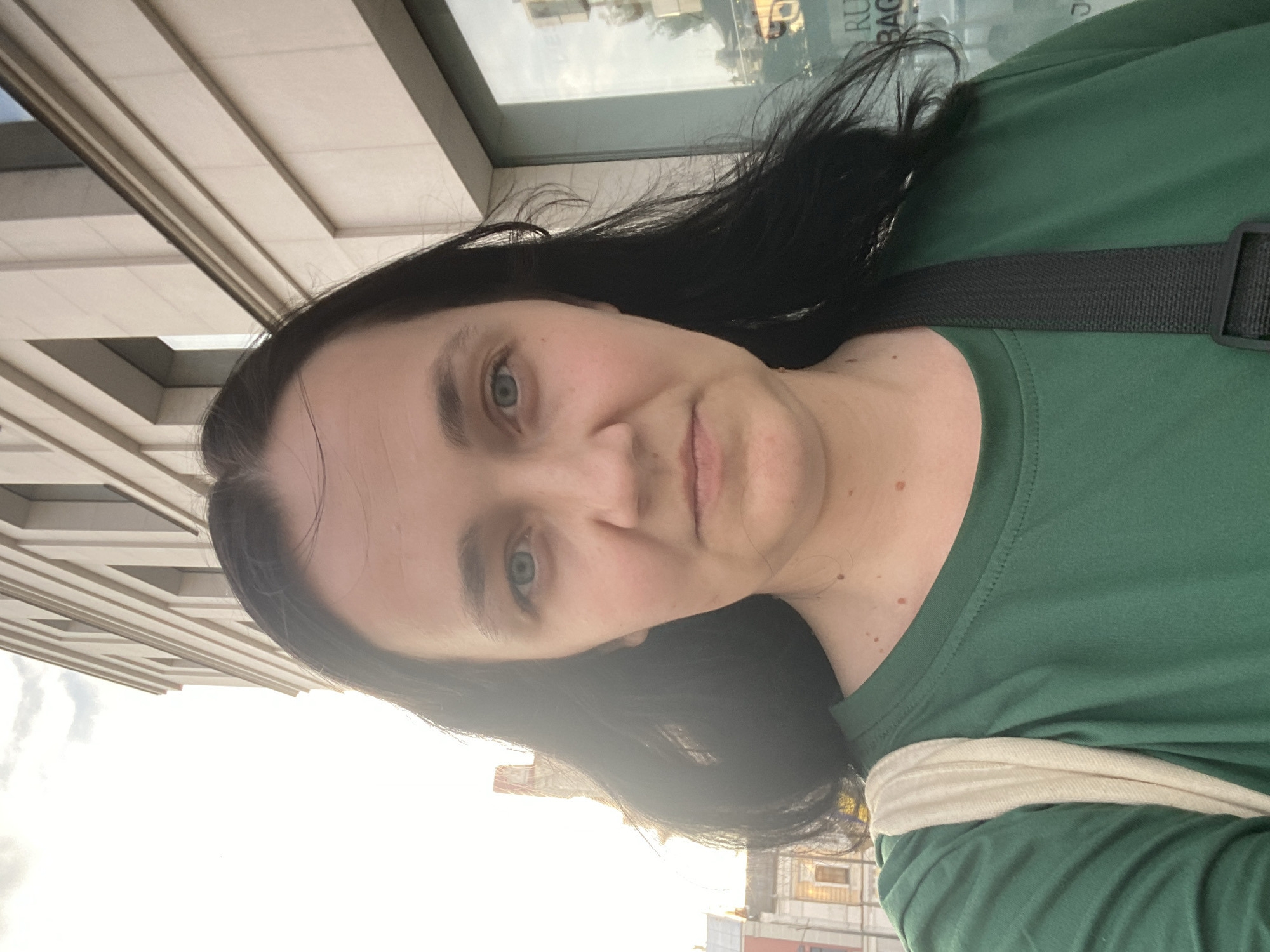 A woman with dark hair and blue eyes is taking a selfie outdoors. She is wearing a green t-shirt and a beige strap from a bag is visible over her shoulder. Behind her, a building with modern architecture and windows is visible, along with a cloudy sky. The context suggests an urban setting, potentially in an area with a mix of old and new architecture.
