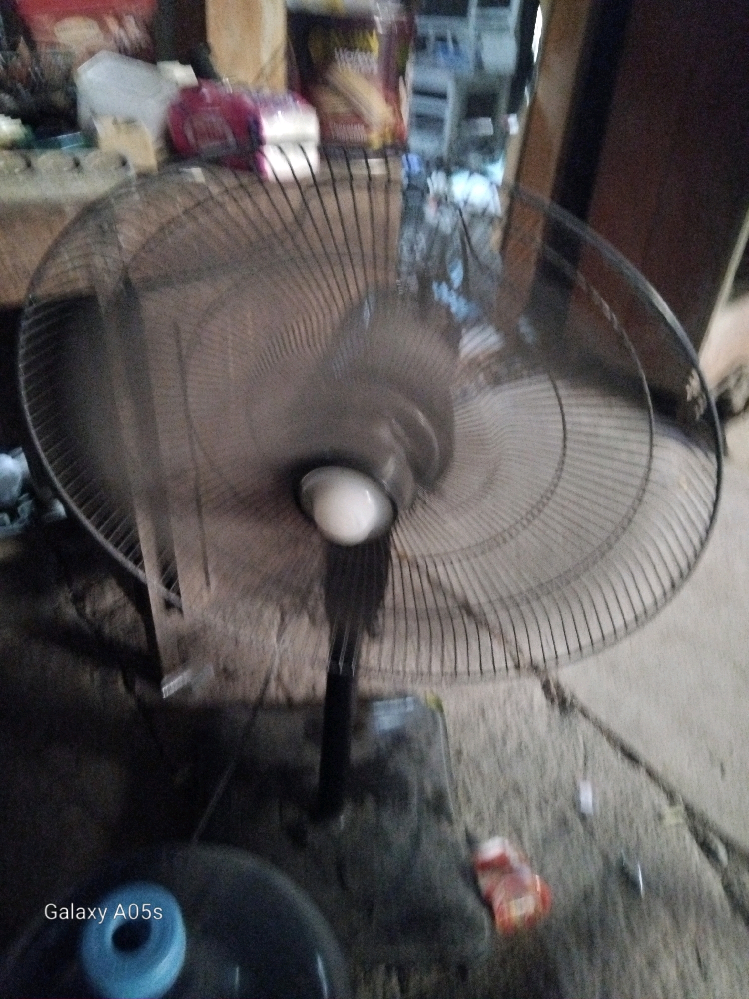 A floor fan is positioned in a room, with its oscillating head blurred due to motion. In the background, shelves hold various items, including what appear to be food products. The floor of the room is made of concrete. The image includes a watermark in the bottom left corner that reads "Galaxy A05s". The location is specified as Karangayung, Indonesia.