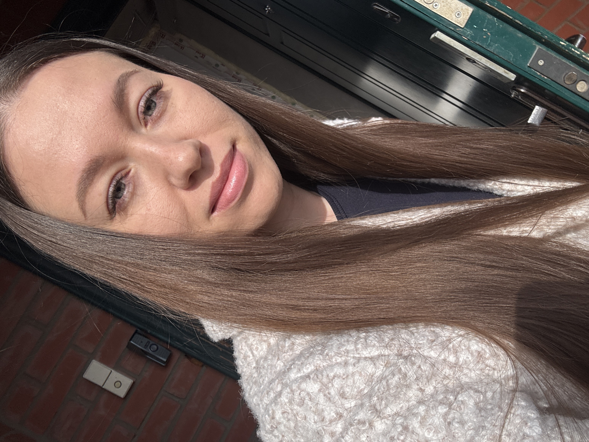 A woman with long brown hair poses for a selfie outdoors. She is wearing a light-colored sweater. Behind her is a dark green door with silver hardware. To the left of the door is a brick wall with a white rectangular object and a black square object attached to it. It is difficult to ascertain the location, or if the location is in the city of Ферма, Russia, based on the photograph alone.