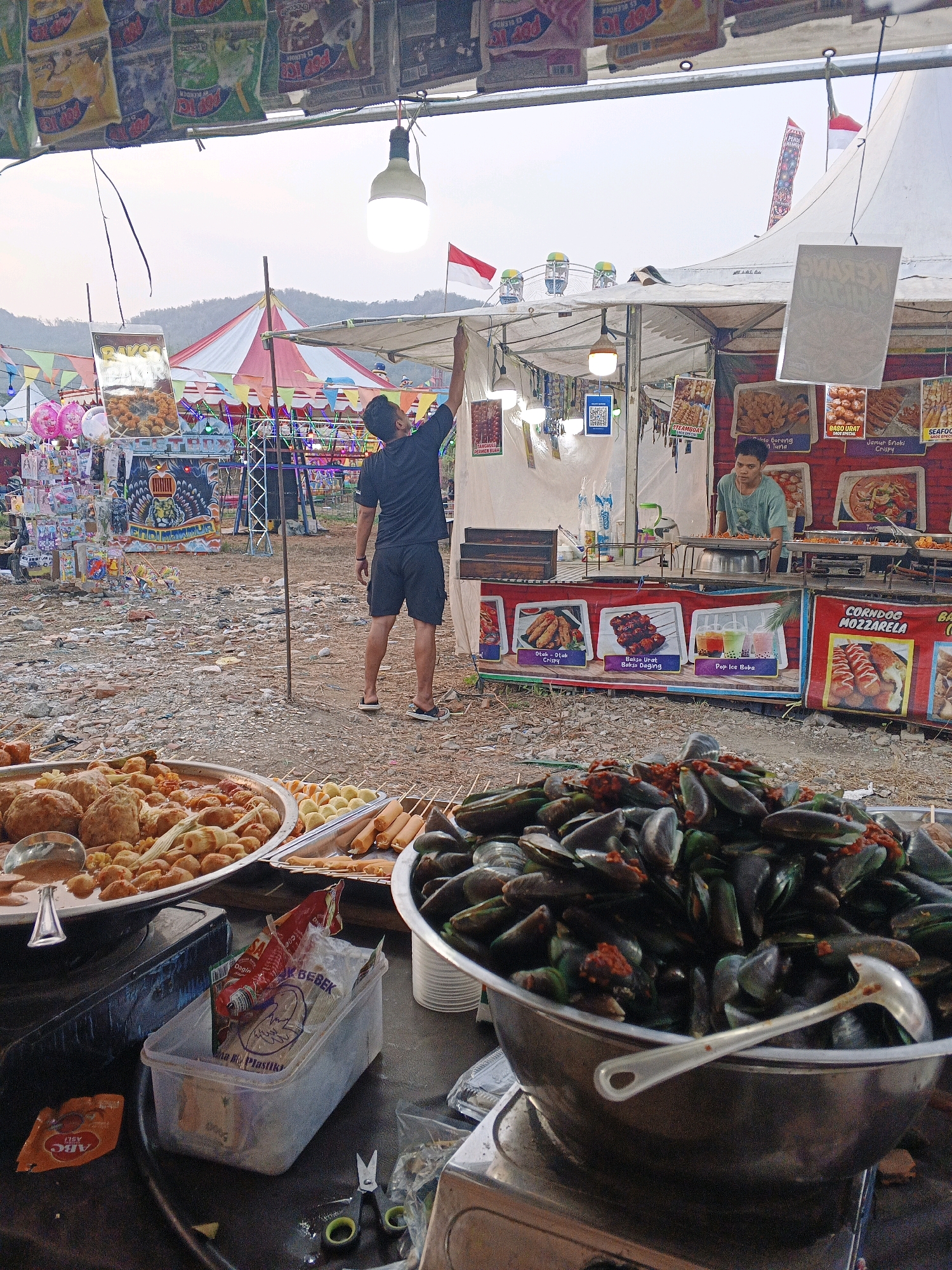 The image depicts an outdoor food stall at what appears to be a carnival or festival in Sukabumi, Indonesia. The foreground is dominated by a large metal bowl overflowing with mussels in a red sauce, suggesting they are a prepared dish. To the left, a metal tray holds an assortment of fried food items, including what look like meatballs and fish cakes, along with skewers of sausages and other small fried items. In the middle ground, a man in a black t-shirt and shorts is reaching up to adjust a light bulb hanging from a makeshift stall structure. Behind him, a food stall operator in a green t-shirt is standing and attending to a cooking station. The stall features various food advertisements displaying images of fried foods and drinks. The background reveals a colorful tent and Ferris wheel, typical of a fairground. The ground is unpaved, covered in dirt and debris, indicating an outdoor, possibly temporary, setup. The overall atmosphere suggests a bustling evening market or festival event.