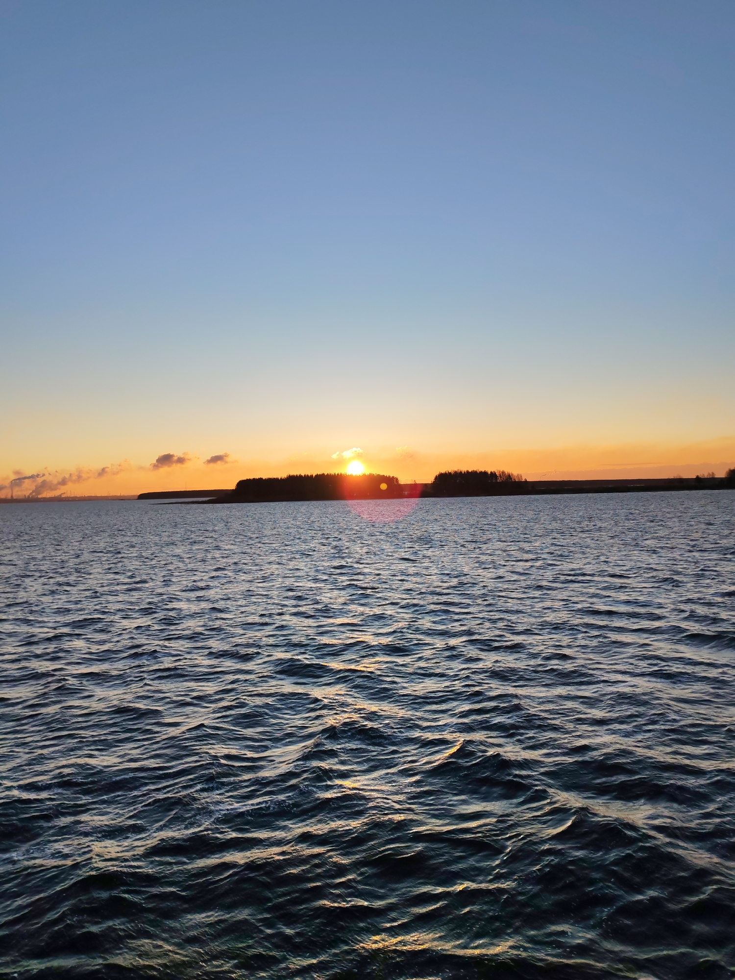 The image shows a body of water with small waves. In the distance, there is a landmass with trees and a hint of buildings or structures on the left side. The sun is setting on the horizon, creating a warm orange glow in the sky. The sky transitions from blue above to orange near the horizon. The location is in the city of Unknown, Unknown.
