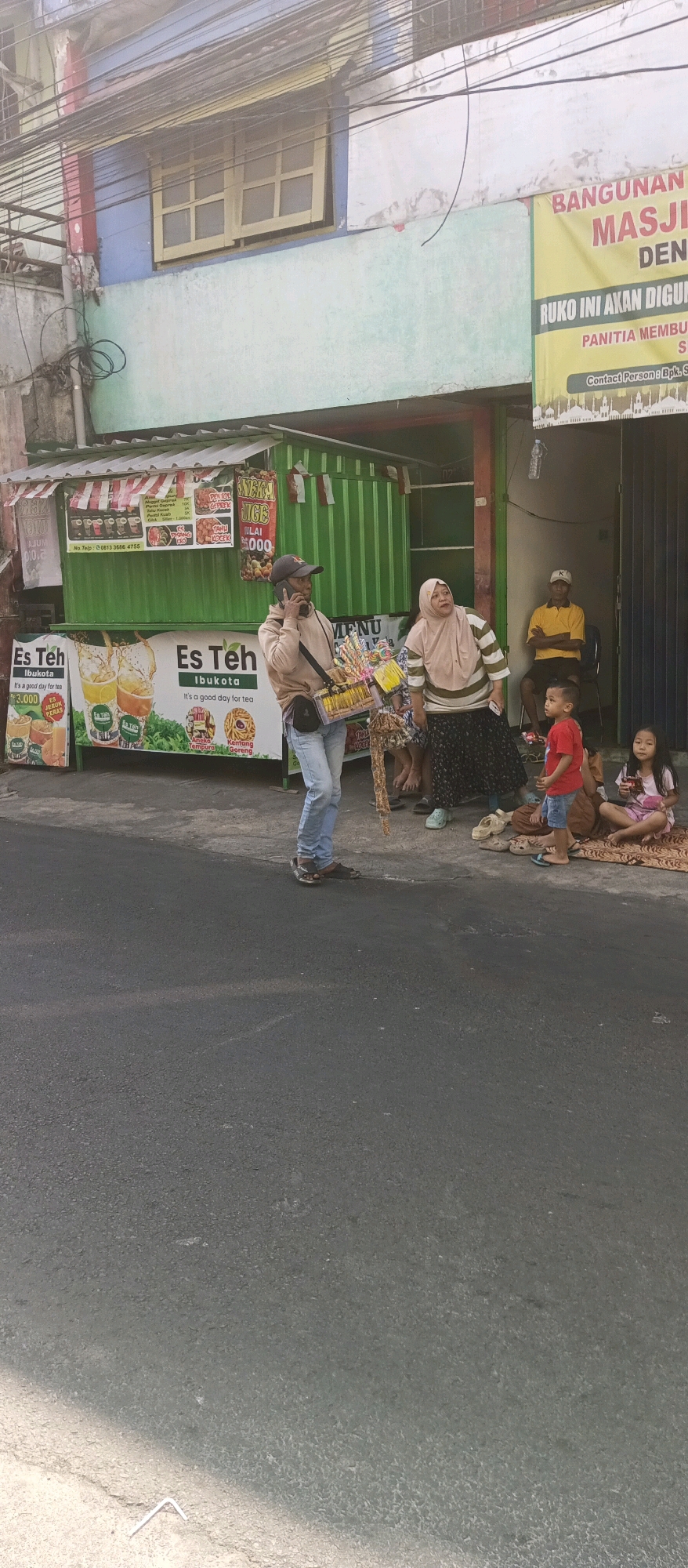 In the city of Malang, Indonesia, a street scene unfolds with a focus on a roadside stall selling "Es Teh" (iced tea). A man, wearing a brown hoodie, blue jeans, and a cap, is speaking on his phone while carrying a basket of assorted candies and snacks. He stands near the stall, which is painted green with white and red striped awnings. Prominent signage on the stall advertises "Es Teh Ibukota" with images of iced tea drinks and snacks like "Kentang Goreng" (fried potatoes).
Next to the stall, a woman in a pink hijab and a patterned skirt looks up and to the right, possibly in conversation with someone off-camera. A group of children are seated on the ground to the right. One young boy in a red shirt and blue shorts sits with his legs crossed, holding a toy. A young girl in a pink dress sits beside him, looking at something in her hands. Another person, partially visible, is seated behind them.
In the background, a yellow banner reads "BANGUNAN MASJID DENGAN..." (Mosque Building With...). A man in a yellow shirt and dark pants is seated in a doorway to the right of the banner, with his arms crossed. The environment suggests a commercial street with buildings, a road, and a sidewalk. Overhead, electrical wires crisscross the scene.