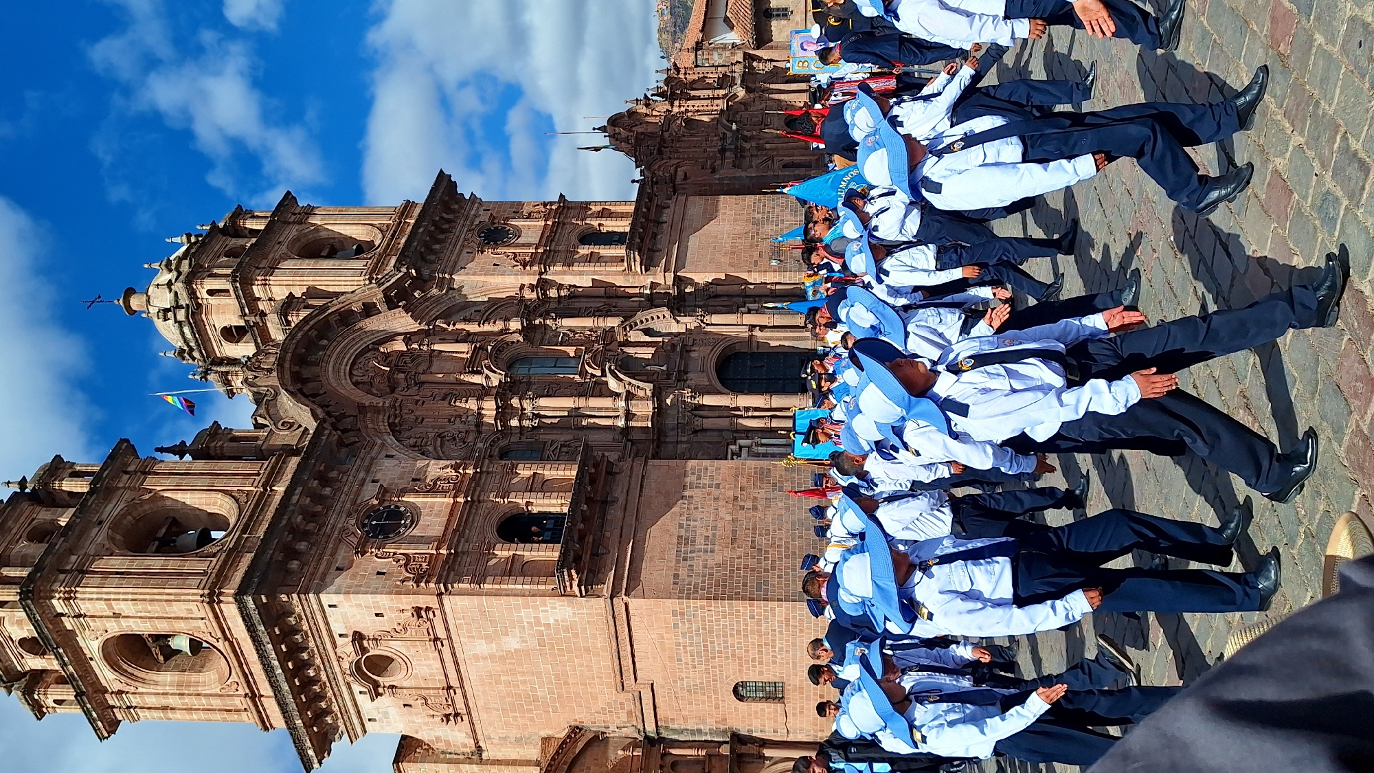 This image captures a procession in the Plaza de Armas in Cuzco, Peru, in front of the Cusco Cathedral. A group of young people, primarily boys, are marching in uniform. They are wearing white long-sleeved shirts, dark ties, dark pants, and matching blue hats with wide brims. Some are also wearing dark jackets or vests over their shirts. They appear to be part of an academic or cadet program, judging by their organized formation and uniforms.
Behind the marching group, the magnificent facade of the Cusco Cathedral dominates the background. The cathedral, a historic colonial-era church, is constructed of reddish-brown stone and features intricate architectural details, including arched doorways, ornate carvings, and towering bell towers. A rainbow flag is visible flying from one of the cathedral's spires, adding a touch of color to the scene. The sky above is a bright blue with scattered white clouds, suggesting a clear and sunny day. The ground is paved with cobblestones, typical of historic city squares. Other individuals, presumably spectators or participants, are visible in the background, some holding flags or banners. The overall atmosphere appears to be one of a formal event or parade.