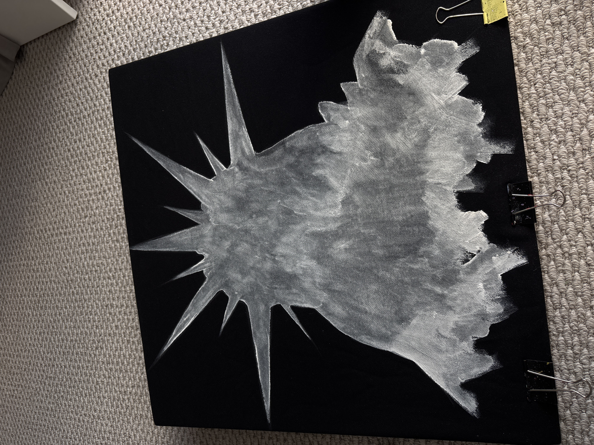 A canvas painting on a textured gray carpet shows a grayscale design. The design features a central area with sharp, star-like points extending outward. To the right of this central area is a blotchy, cloudy shape. The canvas is black. Yellow and black metal clips are attached to the right side of the canvas. A portion of a white object is visible at the top left corner of the image.