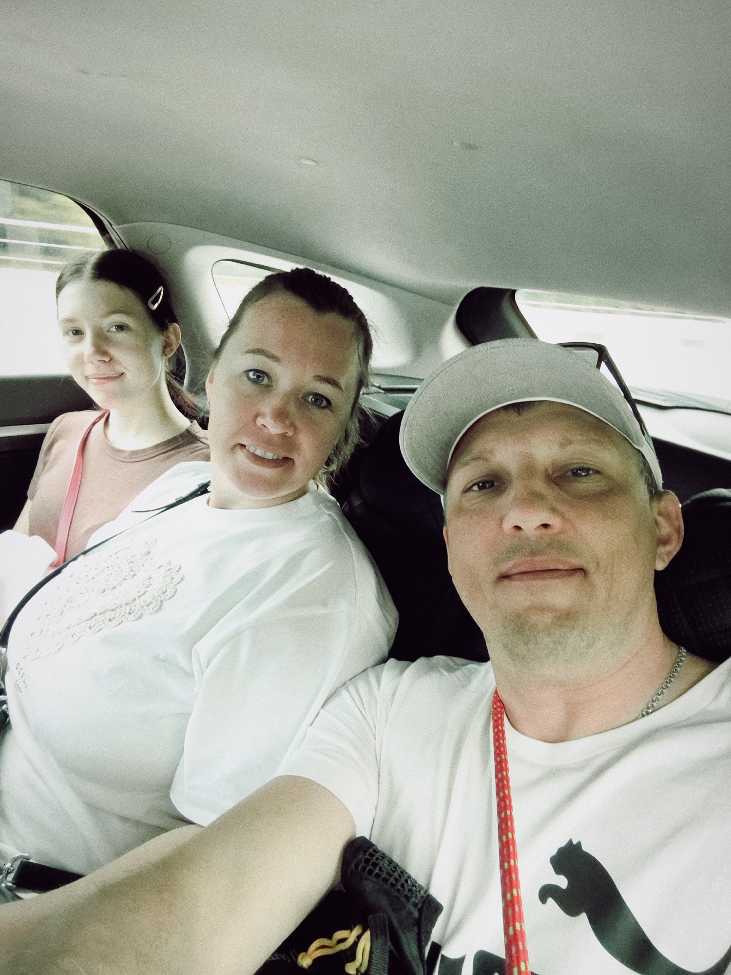 The image shows three people, two women and one man, sitting in the back seat of a car. The woman in the foreground on the right is wearing a white t-shirt and a grey baseball cap. She is smiling and looking at the camera. The woman next to her, in the middle, is wearing a white t-shirt with lace detailing on the chest. She is also smiling and looking at the camera. The youngest woman, on the left, is wearing a brown t-shirt and has her hair pulled back with a clip. She is also smiling and looking towards the camera. The interior of the car is visible, with the headrests and ceiling of the car in the background. The lighting suggests it is daytime. The individuals appear to be on a journey together. There is a visible Puma logo on the man's t-shirt.