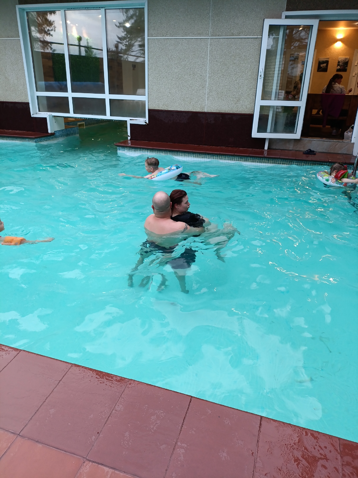 The image depicts an indoor or semi-enclosed swimming pool area in Chaikovsky, Russia, observed from a low angle. The pool contains light blue, rippled water. In the center of the frame, an adult bald man, seen from the back, embraces an adult woman facing right. Further back in the pool, a child in a blue and white inflatable ring is visible, facing away. On the far right, another child wears a red and green inflatable ring and vest. A third person, possibly a child, with an orange arm float, is partially visible on the far left. The pool is bordered by reddish-brown tiles, with a narrow band of blue and white mosaic tiles at the waterline. Above the pool, a dark brown ledge supports a textured, light-colored wall. Two large openings are present: a horizontal multi-paned window on the left, angled upwards, showing reflections of trees and an outdoor area; and an open, white-framed door on the right, leading into an illuminated interior room. Inside this room, a woman with dark hair in a ponytail and a striped shirt is seated at a wooden table. A pair of dark sandals rests near the open doorway on the right.