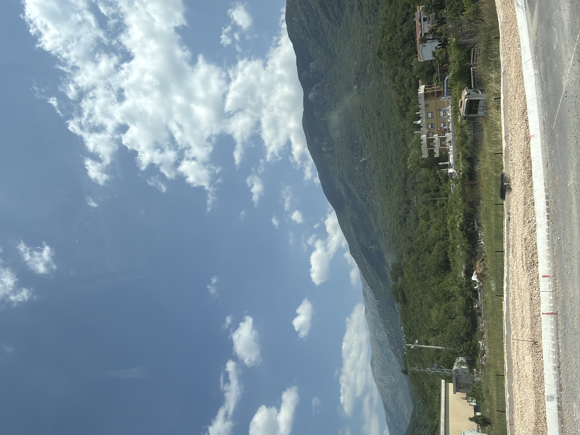 The image depicts a scenic landscape in Kovačko Polje, Montenegro, under a bright blue sky with scattered white clouds. In the foreground, a road with a gravel embankment and a concrete curb is visible. To the left, a light-colored building is partially shown. In the middle ground, a grassy area contains some scattered debris, including what appears to be discarded chairs and a pile of white material. A tall metal tower stands next to a smaller concrete structure. Further back, several buildings are situated on a hillside, including a multi-story building with a light yellow facade and a white tiled roof, and a white building with an orange-tiled roof. The background is dominated by a lush green, forested mountain range. The overall scene suggests a rural or semi-rural environment with some ongoing construction or development.