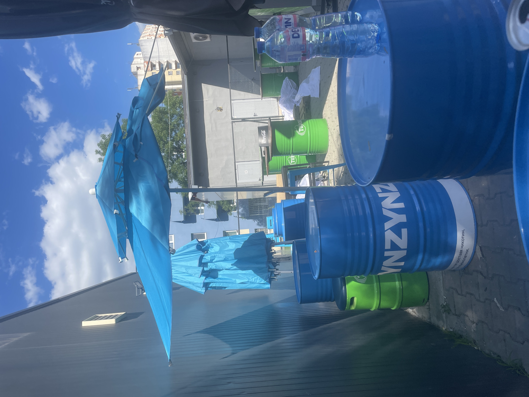 Here is a forensic description of the image:
The image depicts an outdoor setting in Sofia, Bulgaria. There are numerous repurposed oil drums, painted in vibrant blue and green, used as tables or seating. A row of blue umbrellas offers shade. In the background, an industrial-looking building and a partial view of an adjacent building can be seen, with a clear, blue sky filled with scattered clouds above. Two water bottles are visible on top of one of the blue barrels. Some green barrels have Coca-Cola branding. A white cloth is placed on a small table. There are markings on the blue barrels that include the word "YNZYNZ" and a short description in Cyrillic. The ground consists of paved stones. A partially visible black structure (possibly an umbrella or awning) is located in the upper part of the frame.