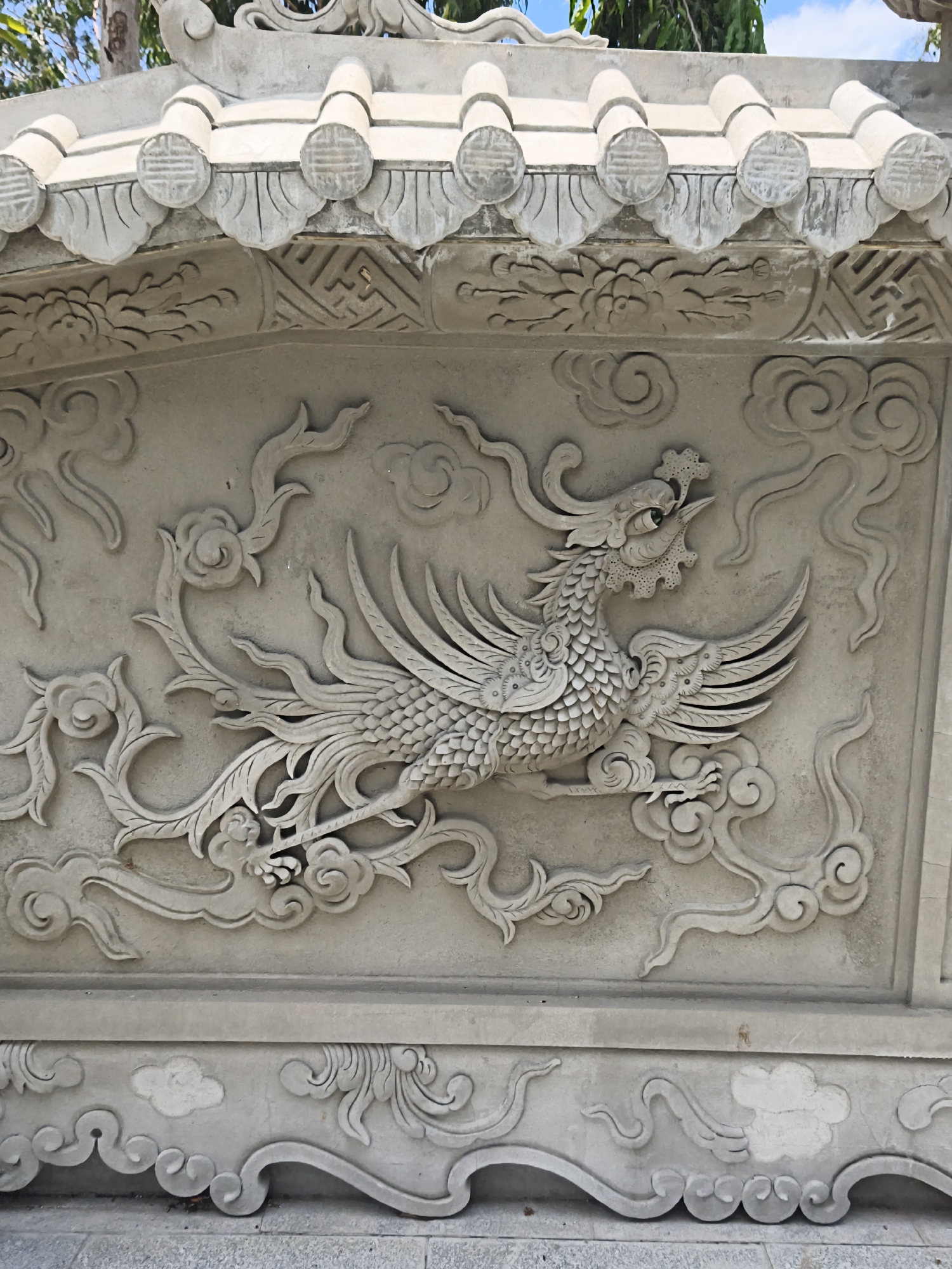 A close-up, low-angle outdoor shot shows a relief carving on a light-colored stone wall. The carving features a stylized bird, possibly a phoenix, with intricate feather details and a scaly body. The bird is depicted in profile, facing right, with its beak slightly open. It is surrounded by swirling cloud motifs and floral elements. Above the bird carving is a decorative frieze with patterns and what appear to be stylized roof tiles. The overall style suggests East Asian architecture or artistry. The stone appears weathered, with some areas showing discoloration and minor damage. The background is blurred but hints at foliage and a blue sky.