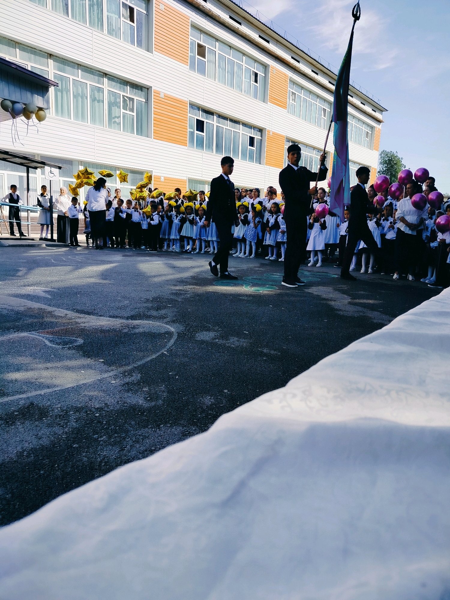 The image depicts an outdoor event at a school in Kölkent, Kazakhstan. The foreground shows a textured, dark asphalt ground with white chalk markings, and a large white draped fabric covering part of the bottom right. In the mid-ground, a group of students, dressed in formal attire including suits and dresses, are gathered, some holding balloons. A few adults are also present, interacting with the students. A person is holding a flag pole with a colorful flag. In the background, a modern school building with multiple windows and an orange accent on its facade is visible. The sky is clear and blue. The overall atmosphere suggests a school celebration or ceremony.