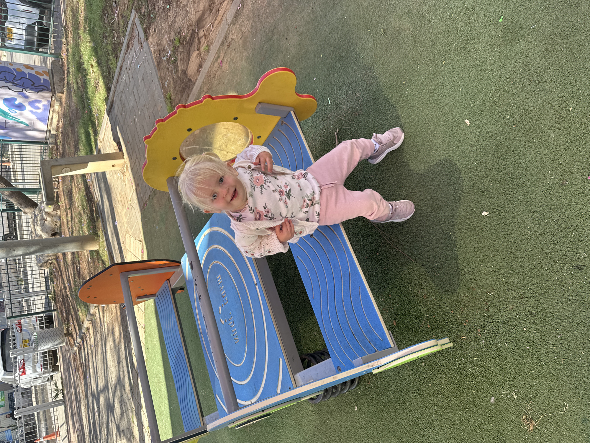 A young child with blonde hair and blue eyes is captured smiling directly at the camera while sitting on a colorful playground spring rider. The child is dressed in a white long-sleeved top with a pink floral pattern, a light-colored cardigan, pink sweatpants, and grey sneakers.
The playground equipment features blue wavy platforms, a yellow sun-like top, and red accents, with a black spring visible underneath. The ground beneath the equipment is green artificial turf. In the background, to the left (if the image were upright), a metal fence, a paved path, trees, and a white car with an Israeli license plate (64-287-21) are visible. A concrete structure, possibly a light post or water fountain, stands near the fence.
The text "עולם הילד מבית מותק" (Olam HaYeled MiBayit Motek), translating to "The Child's World from Motek House," is inscribed in Hebrew on the blue section of the playground equipment.
The scene takes place outdoors on a bright, sunny day, likely in the late morning or early afternoon, evidenced by the distinct shadows. The child's happy expression conveys enjoyment. The setting is a public playground, consistent with the Petah Tikva, Israel, location, which is further supported by the visible Hebrew text and Israeli license plate.