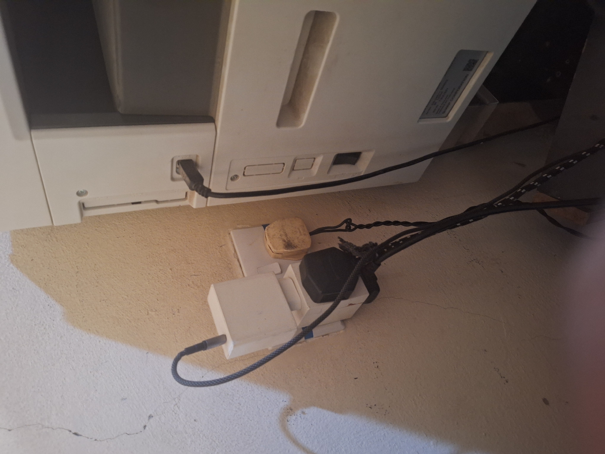 The image shows a wall with a power strip plugged into a wall socket. Multiple adapters are plugged into the power strip, and various cables are connected to them. A printer is visible on the right side of the image, with a cable plugged into it. The wall appears to be textured and painted in a light color, with some cracks visible. The overall environment suggests a residential or office setting in Jalingo, Nigeria, possibly an area with limited electrical infrastructure.