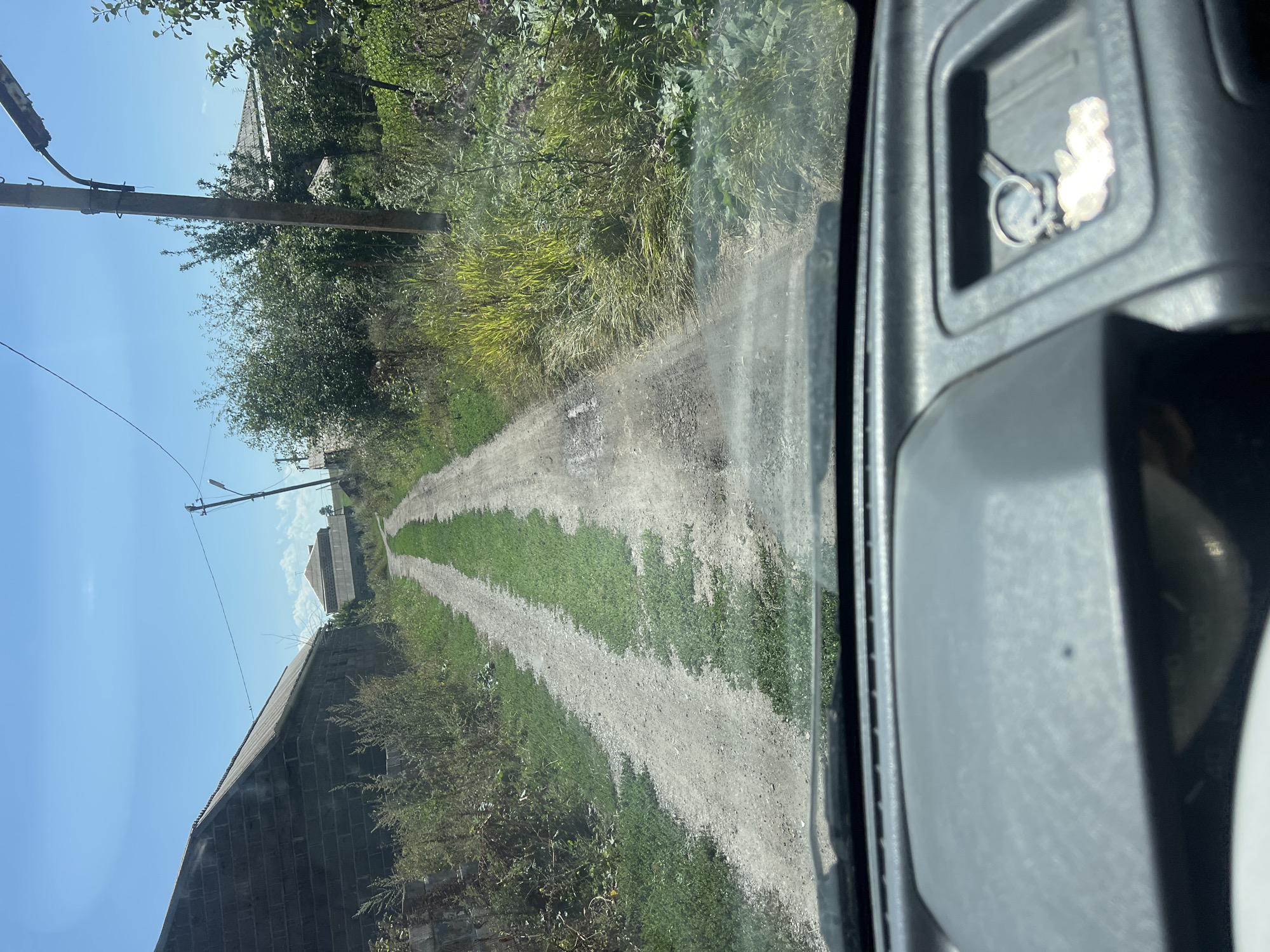 The image shows a view from inside a vehicle driving down a dirt road in Khando, Georgia. The road is flanked by overgrown vegetation and appears to be in a rural or semi-rural area. On the left side of the road, there is a dark brick building, possibly a house or a barn. Further down the road, there are other buildings visible, suggesting a small settlement. On the right side, there is a utility pole with a light fixture and dense foliage. The sky is clear and blue, indicating a sunny day. The interior of the vehicle is visible in the foreground, with part of the dashboard and a small compartment holding keys and a keychain with a charm.