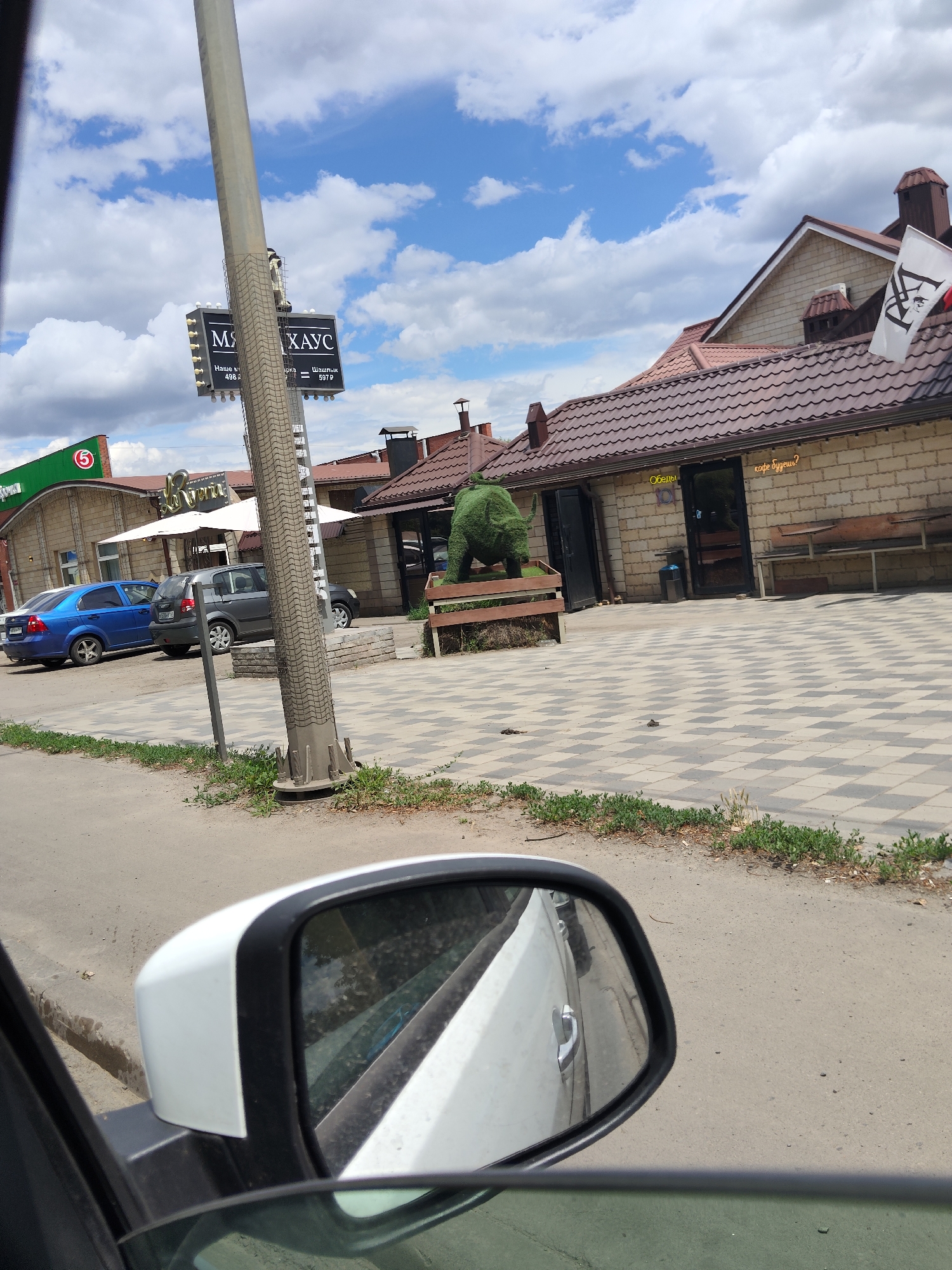 The image captures a view from inside a car looking out at a street scene in Temernitskiy, Russia. A white side-view mirror is prominently featured in the foreground. Beyond the mirror, a paved area with a checkered pattern is visible, leading to a building constructed with light-colored bricks. The building has a brown, tiled roof and features several windows and doors. A green, topiary-like sculpture resembling an animal is positioned on a wooden bench in front of the building. Several cars are parked on the side of the road. A tall pole with signage is also visible, displaying text that appears to be in Cyrillic script. The sky above is blue with scattered white clouds, suggesting it is daytime and fair weather. A white flag with a black emblem is attached to the roof of the building.