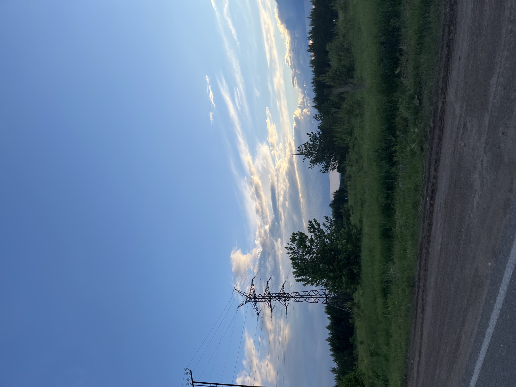 The image depicts an outdoor scene with a focus on a power line tower. The tower is situated in a grassy area adjacent to a road. The sky is visible above, displaying streaks of clouds. The presence of a paved road with a white line marking suggests an urban or suburban environment. Considering the question's reference to the city of Chaikovsky, Russia, it is reasonable to assume this scene is located in or near that city. The image does not contain people, but it shows evidence of infrastructure (the power line) and transportation (the road) within a natural environment. The interaction between these elements creates an image of a place where human activity and nature coexist.