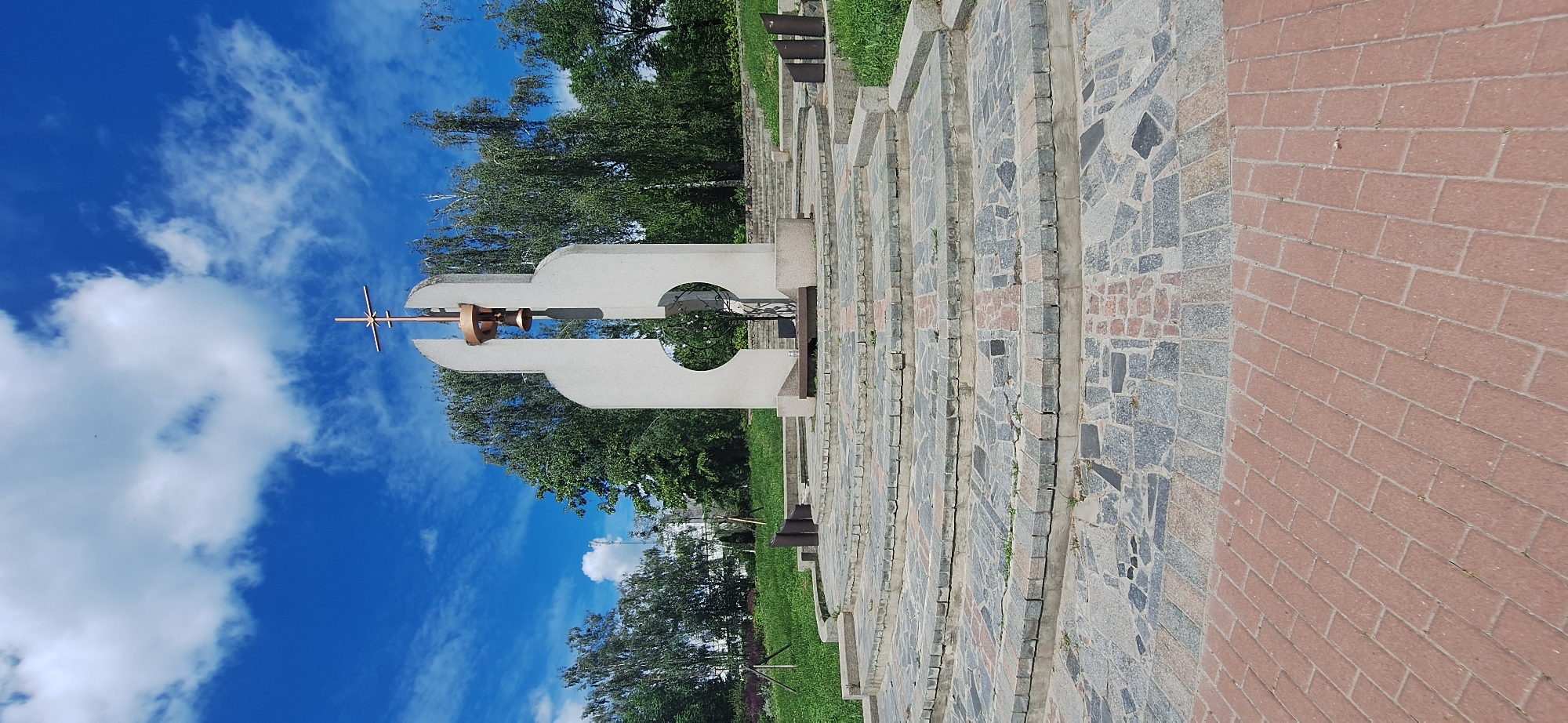 The image depicts a monument with a cross and bell, situated in a park-like setting in Bila Tserkva, Ukraine. The monument is made of light-colored concrete and has a modernist design. It is surrounded by tiered stone steps and paved walkways made of red bricks and grey stones. Green grass and trees form the background, with a bright blue sky and white clouds above. The scene is peaceful and appears to be during the daytime. There are no people visible in the image. The monument is likely a place of historical or religious significance.