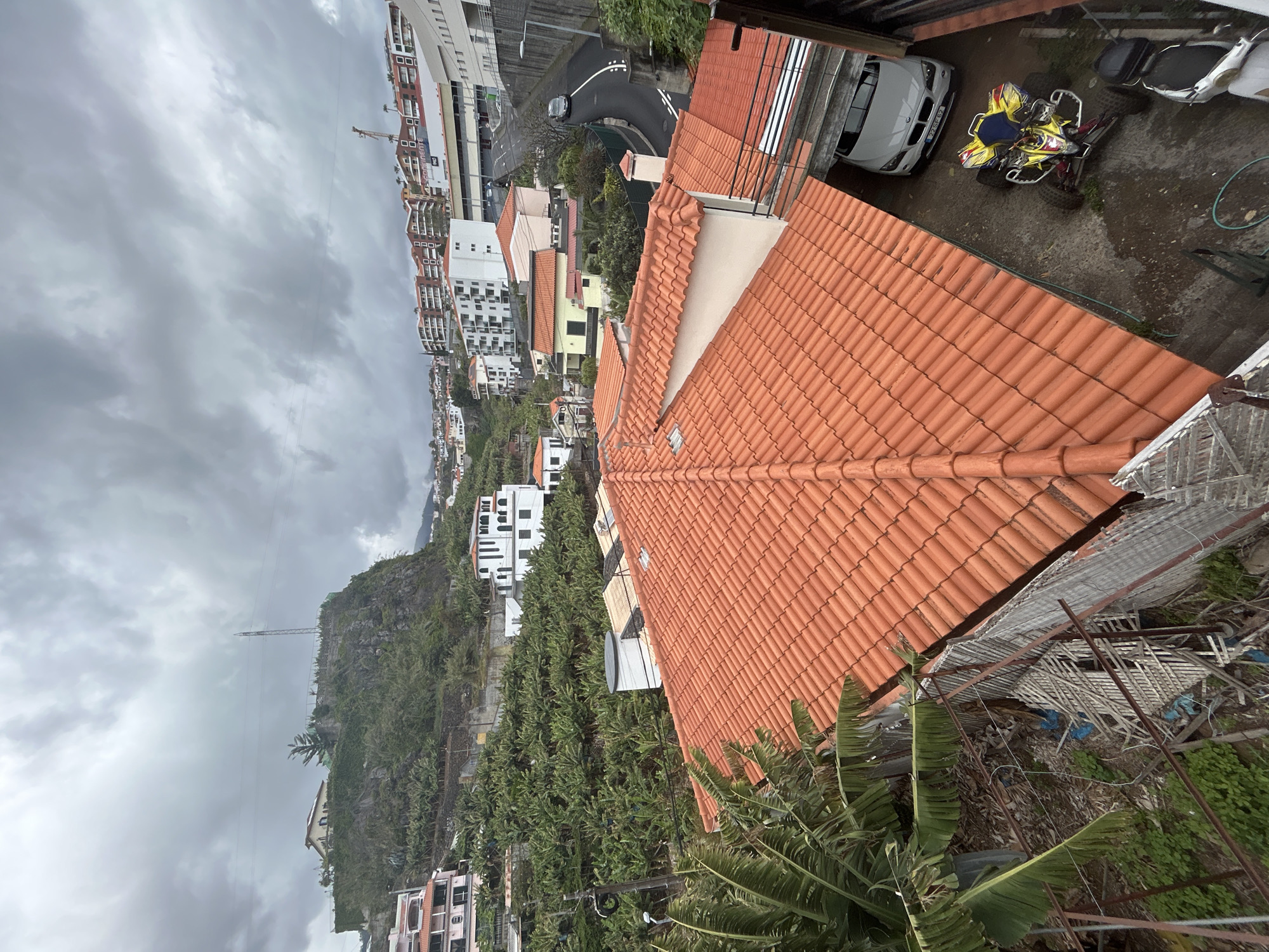 A high-angle shot of Câmara de Lobos, Portugal, showcases a mix of urban and natural elements. The architecture includes tiled rooftops, multi-story buildings, and houses nestled among green spaces. The landscape features banana plantations covering hillsides. The sky is overcast with dark, heavy clouds. In the foreground, there is a tiled rooftop and some vegetation. A vehicle and an ATV are parked nearby. Overall, the scene captures the blend of residential structures and verdant, hilly terrain in Câmara de Lobos.