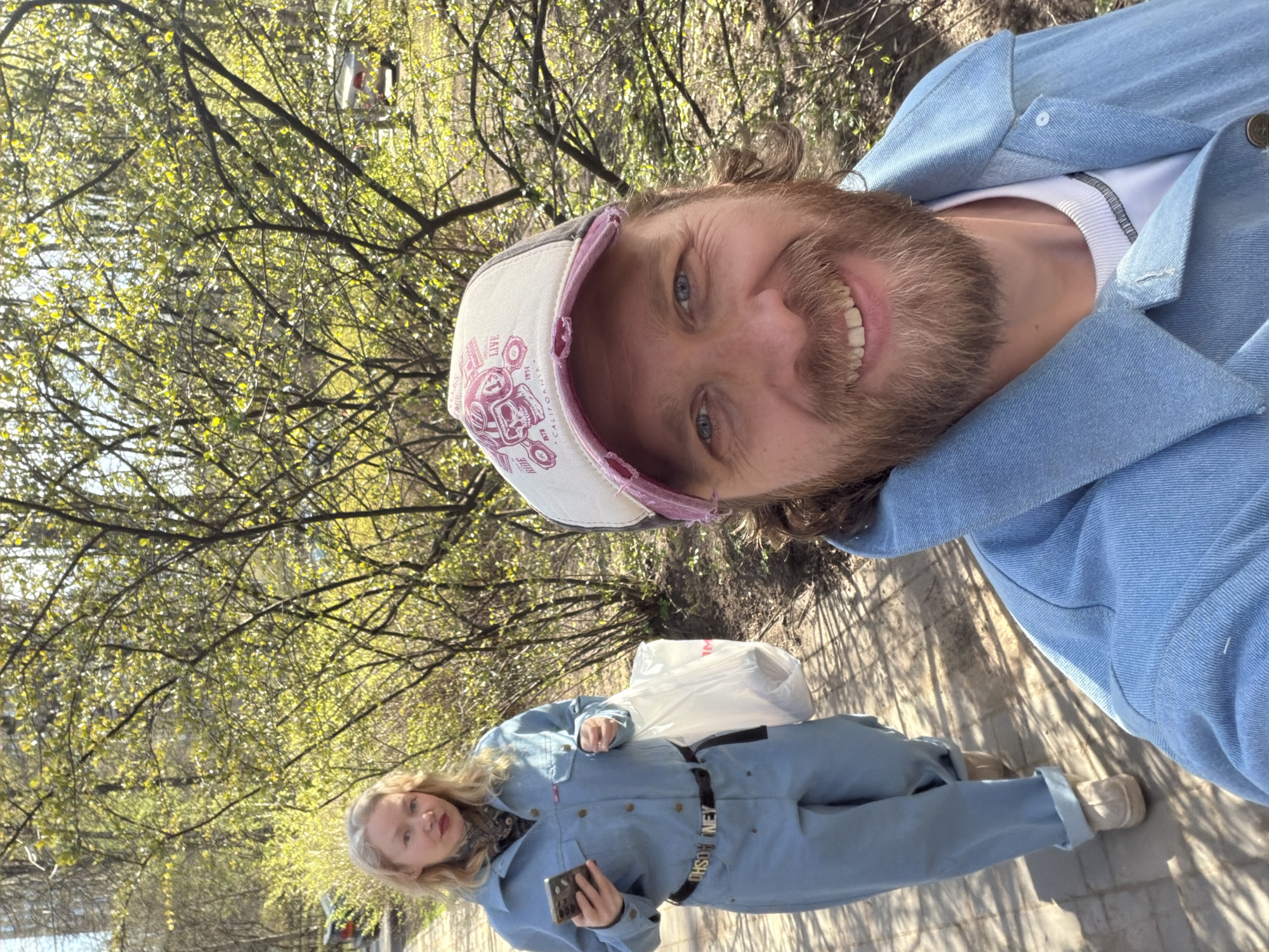 This outdoor image, taken in Saint Petersburg, Russia, features two individuals. In the foreground, positioned to the right, a bearded man with blue eyes is taking a selfie. He is smiling broadly, wearing a light-colored cap, a light blue denim jacket, and a white t-shirt. His cap has some stylized, partially visible text that appears to include "CALIF" and possibly "RIDE."
In the background, standing further down a paved path and slightly to the left, is a woman with blonde hair. She is dressed in a light blue denim jumpsuit, cinched at the waist with a black belt, and light-colored shoes. She holds a smartphone in her hand and looks towards the camera with a neutral expression. Behind her, a white plastic bag is visible on the ground. The black belt on her jumpsuit clearly displays the text "KEN MOSH," which might be part of a brand name.
The setting is a sunny, spring day, with bright daylight casting shadows on the paved path. The background is dominated by lush trees with numerous light green leaves, indicating early spring foliage. The overall atmosphere appears casual and relaxed, suggesting a stroll or outing in a park or tree-lined area.