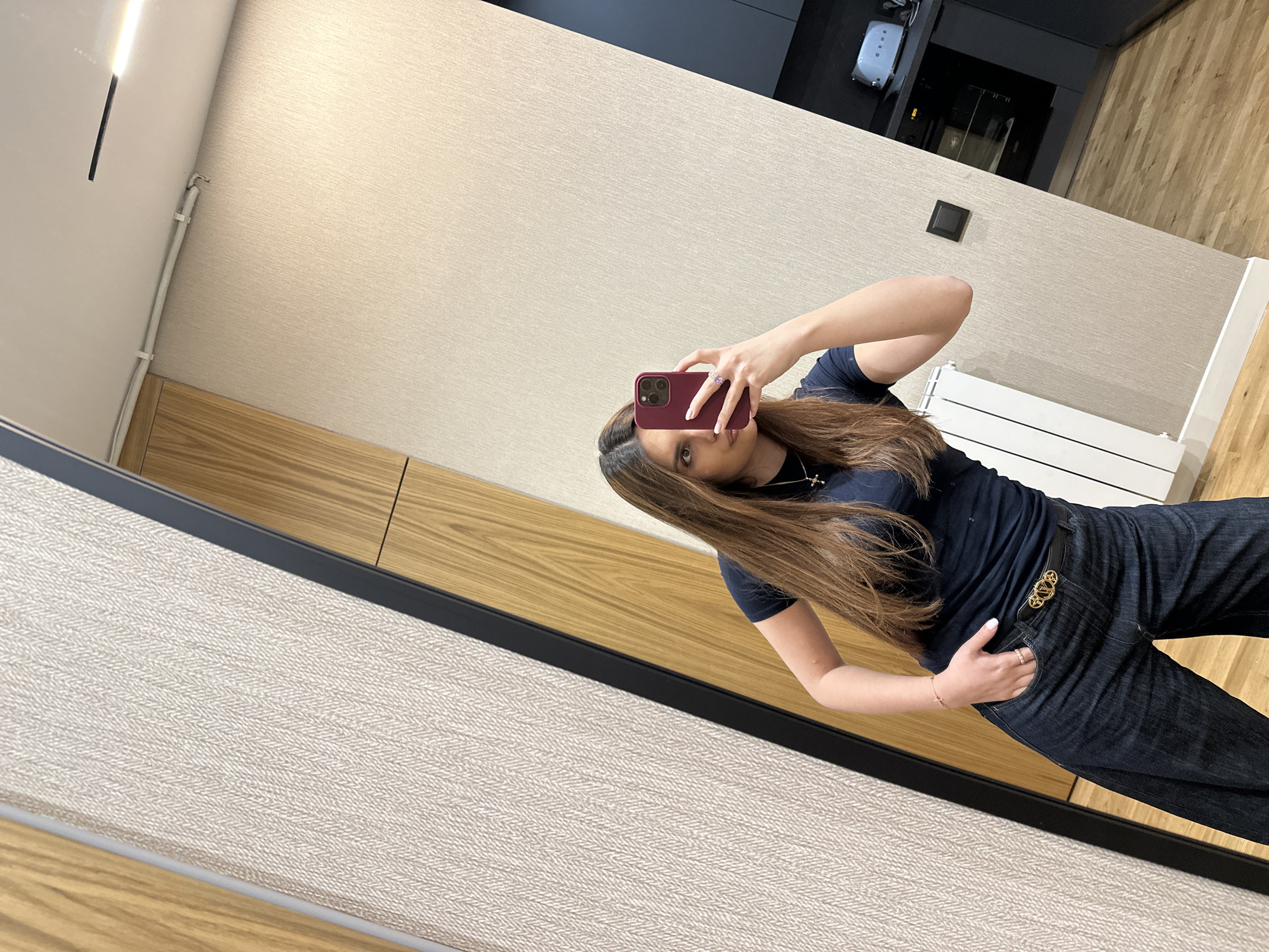 A woman is taking a selfie in a mirror in what appears to be a retail space. She is holding a maroon-colored smartphone with her right hand, partially obscuring her face. The woman has long, brown hair and is wearing a dark blue t-shirt and dark jeans with a distinctive gold-colored belt buckle. She has jewelry on her fingers and wrists. The background consists of a wall with alternating sections of beige textured material and light wood paneling. A black frame surrounds the mirror. In the upper part of the mirror's reflection, there is a glimpse of what looks like an elevator or service area. The location is indeterminate, but the request specified the city of Yerevan, Armenia.