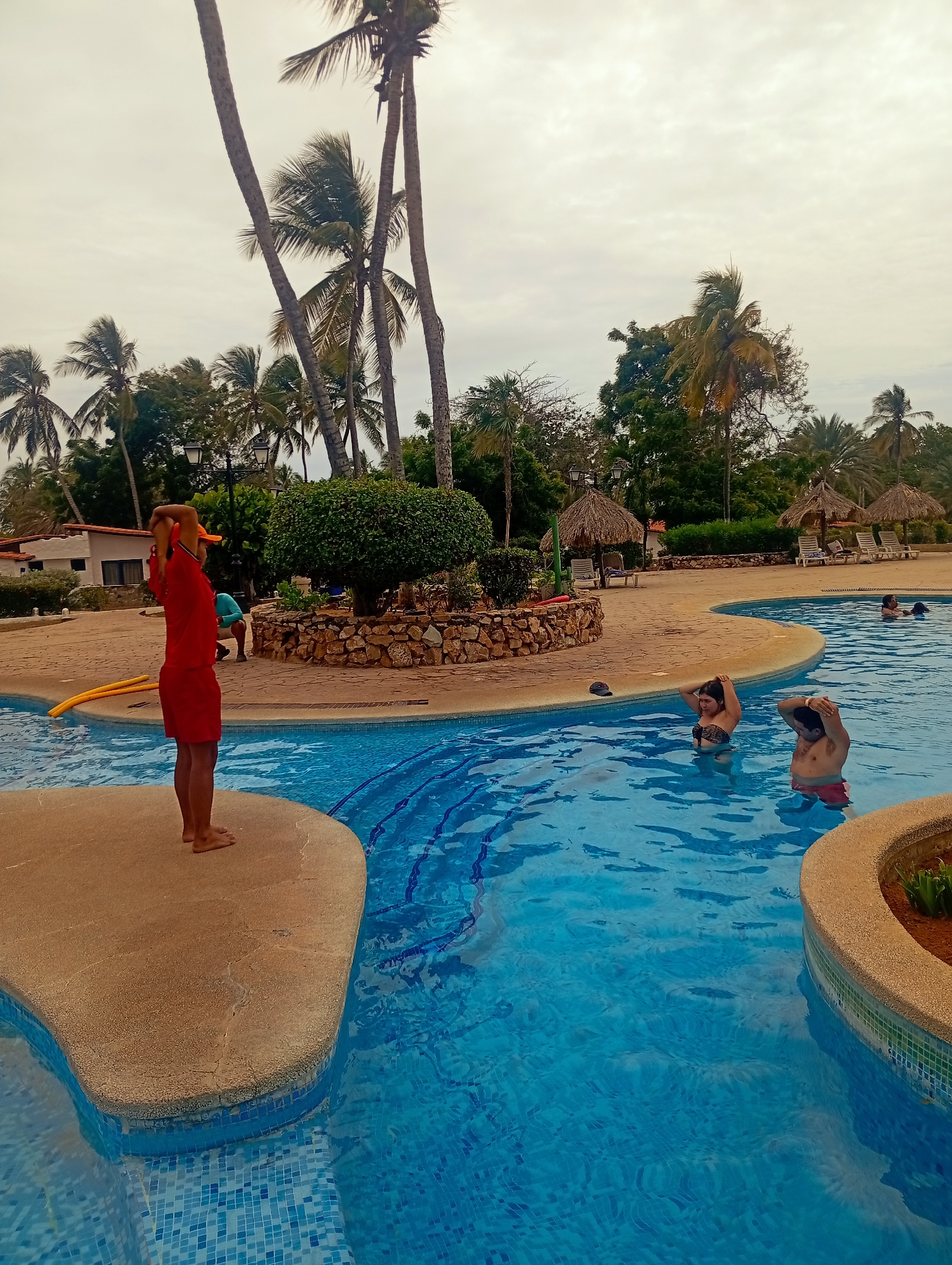 This image captures a vibrant scene at a tropical resort swimming pool, likely located in Playa El Agua, Venezuela. The setting is characterized by lush tropical vegetation, dominated by numerous tall palm trees that punctuate the overcast sky.
The central feature is a large, irregularly shaped swimming pool with a tiled blue bottom, showing subtle patterns from deeper blue lines. The pool is bordered by light brown, textured paving, which also forms a small, elevated platform and a circular garden bed with green shrubs. Thatched-roof palapas with lounge chairs are visible in the background, nestled among the foliage, suggesting a leisure or vacation destination.
Several people are present, indicating an ongoing activity. In the foreground, a person stands on the pool platform, wearing a red t-shirt, red shorts, and an orange cap. Their arms are raised over their head, suggesting they are leading an exercise or warm-up session. Yellow pool noodles lie on the pavement nearby.
Inside the pool, a woman wearing a dark bikini top and a man in red swim shorts are partially submerged, both with their hands behind their heads, seemingly mimicking the pose of the person on the platform. Another individual is visible further back in the water. In the mid-ground, near the garden bed, another person in a light blue top is bending over.
The time of day appears to be mid-day or afternoon, under an overcast sky, which provides soft, diffused lighting without harsh shadows. The overall mood is relaxed and active, typical of a resort setting. No visible text is present in the image.
