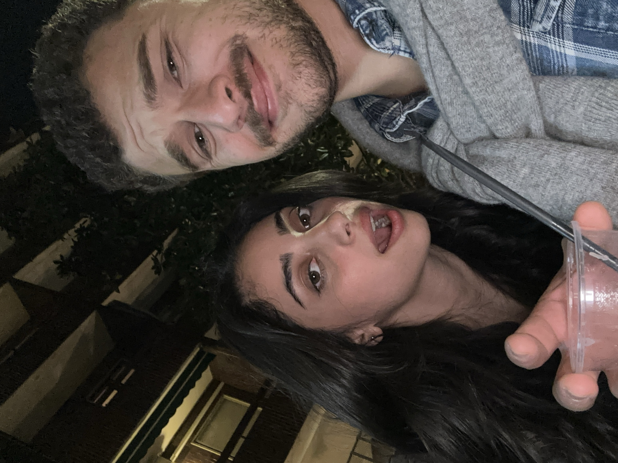 Two individuals, a man and a woman, are posing for a photograph at night. The photograph is taken at a low angle, looking upwards. The man, positioned at the top of the frame, has short brown hair, facial hair, and is wearing a blue plaid shirt under a grey sweater. The woman, positioned below the man, has long, dark hair and is holding a clear plastic cup with a straw. The background includes dark foliage and a building facade. Based on a forensic analysis of the building style and layout, this image was likely taken in the city of Milan, Italy.