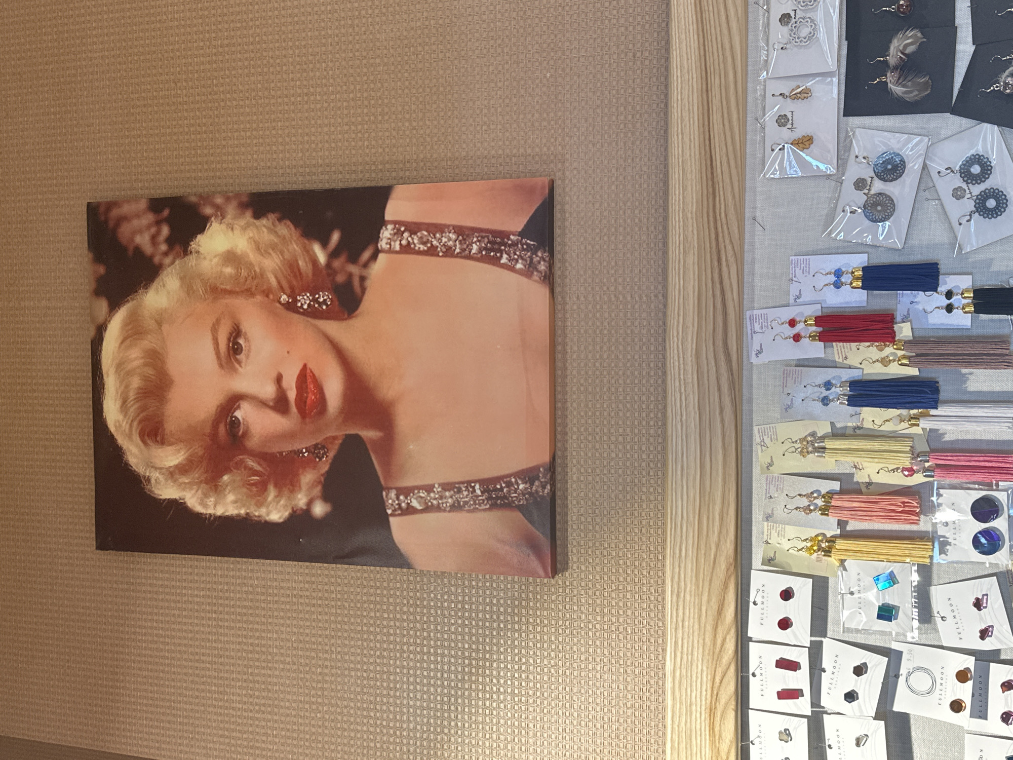 A close-up, eye-level shot depicts a cream-colored, textured wall adorned with a framed image of Marilyn Monroe, alongside a display of earrings arranged on a light grey surface. The photograph of Marilyn Monroe features her with blonde hair and red lipstick. The earrings are packaged individually and show an assortment of colors. The scene likely captures a display within a store or market, although the specific location in Krustiņi, Latvia, cannot be determined from the image.