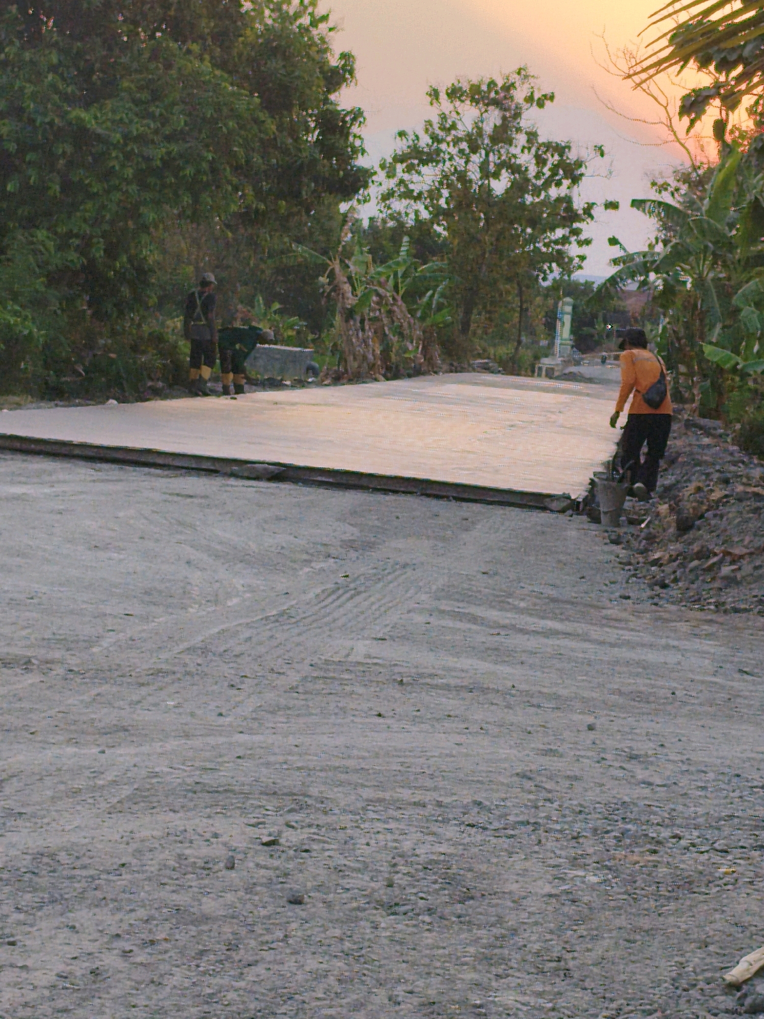 This photograph captures a scene of road construction in Gondangrejo, Indonesia, at sunset. Two individuals are present, both appearing to be workers involved in the construction. One worker, wearing a dark shirt, dark pants, and yellow boots, stands near a wheelbarrow on the left side of the image. Another worker, dressed in an orange long-sleeved shirt and dark pants, is positioned on the right, carrying a bag over their shoulder and standing near a bucket. The construction site features a newly laid, smooth concrete surface, possibly a road extension or repair, bordered by rougher, unfinished ground. Vegetation, including trees and banana plants, flanks the road. The sky is illuminated by the warm hues of sunset. The overall impression is one of ongoing labor amidst a rural or semi-rural setting in Indonesia.
