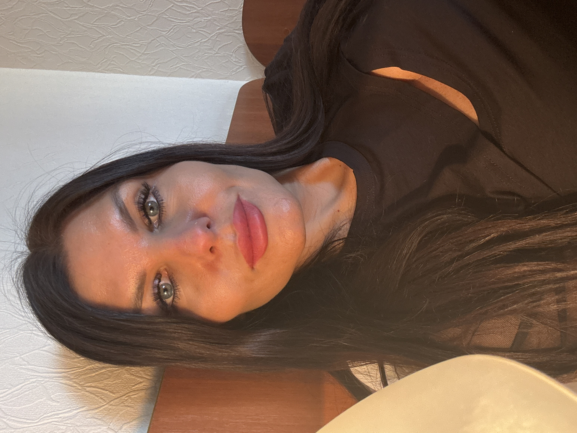 The original image is a rotated close-up portrait of a woman, likely taken indoors in Perm, Russia. The woman, with long, dark hair, features striking blue-green eyes and full lips accentuated by pink lipstick. Her gaze is directed towards the camera with a neutral to slightly serene expression. Her fair skin shows subtle highlights.
She is dressed in a dark, likely black, t-shirt or top with a wide neckline, revealing a portion of her shoulder or collarbone on the right side of the frame.
The background includes a light-colored, textured wall covering or wallpaper visible in the upper left quadrant. A dark wooden surface, possibly a headboard or another piece of furniture, is situated behind her and to her right. A curved, light-colored object, perhaps a lamp or a cushion, is partially visible in the bottom right corner. The lighting appears soft and diffused, suggesting an indoor setting without harsh direct light. No text is visible in the image.