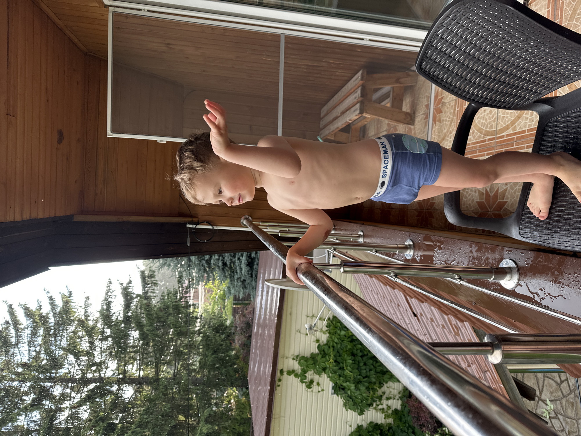 A young boy with blond hair stands on a wooden balcony in Nizhny Novgorod, Russia, wearing blue boxer briefs with a "SPACEMAN" logo. He is looking to the left with his right arm raised. The balcony has a metal railing that is wet, indicating rain. Outside the balcony, trees and houses are visible, and it appears to be raining. A dark plastic chair is placed on the balcony in front of the boy. The balcony floor is made of wood and is also wet. There is a screen door on the right side of the balcony, and a wooden bench can be seen through it.