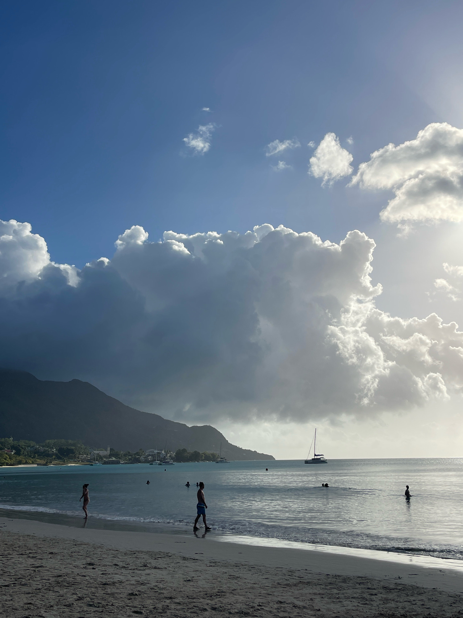 A wide-angle, eye-level shot captures a tropical beach scene on a sunny day with dramatic clouds overhead. The foreground features a sandy beach, with gentle waves lapping at the shore. Two individuals are walking near the water's edge. One, in a light-colored bikini, walks towards the left. The other, in dark swim trunks, walks towards the right.
The middle ground shows a calm, turquoise body of water. Several people are visible in the water, with some appearing to be swimming. A sailboat with a white hull and a dark sail is positioned further out in the sea.
In the background, a lush, green mountainside rises from the coastline. A small town or village with buildings and vegetation is nestled at the foot of the mountain. Sailboats and other vessels are anchored or sailing in the bay.
The sky is a vibrant blue, partially obscured by large, voluminous clouds. Some clouds are bright white, illuminated by the sun, while others are darker and appear to be gathering or approaching. The overall lighting suggests it is late afternoon, with the sun casting a bright glow on the right side of the scene.
The location is identified as Mare Anglaise, Seychelles.