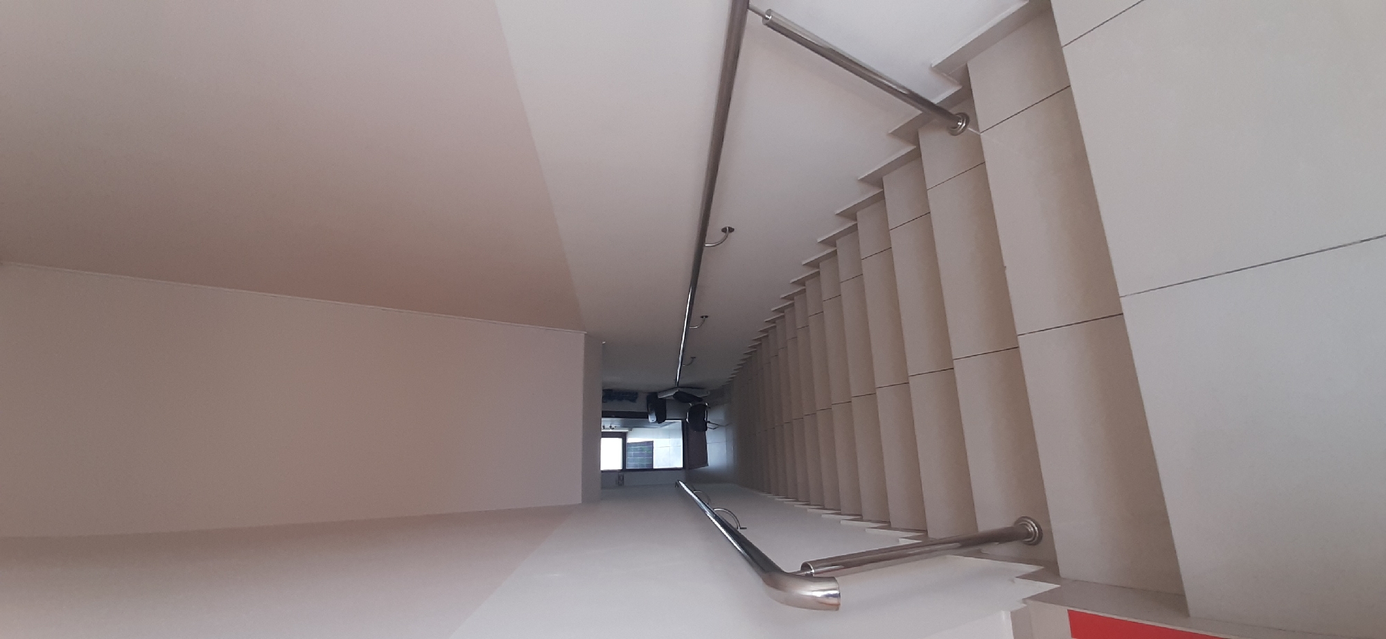 This media file depicts an indoor staircase with a minimalist, modern design, likely located in Yessentuki, Russia. The scene is captured from a high vantage point, looking down into the stairwell. The predominant colors are white and light beige, creating a bright and airy atmosphere.
The staircase features sleek, polished metal handrails that run along the walls, providing a contemporary touch. The walls themselves are adorned with large, rectangular panels in a light beige hue, separated by thin, dark lines, giving them a structured and clean appearance. The ceiling is also white and appears to be a continuous surface.
Further down the stairwell, a glimpse of what appears to be a small room or corridor is visible through an opening. This area is darker than the main stairwell, and a window can be seen, suggesting it might be an external view or another part of the building. The overall impression is one of a well-maintained and aesthetically pleasing interior space, possibly part of a public building, a modern apartment complex, or a commercial establishment. There is no visible text in the image. The lighting suggests it is daytime.