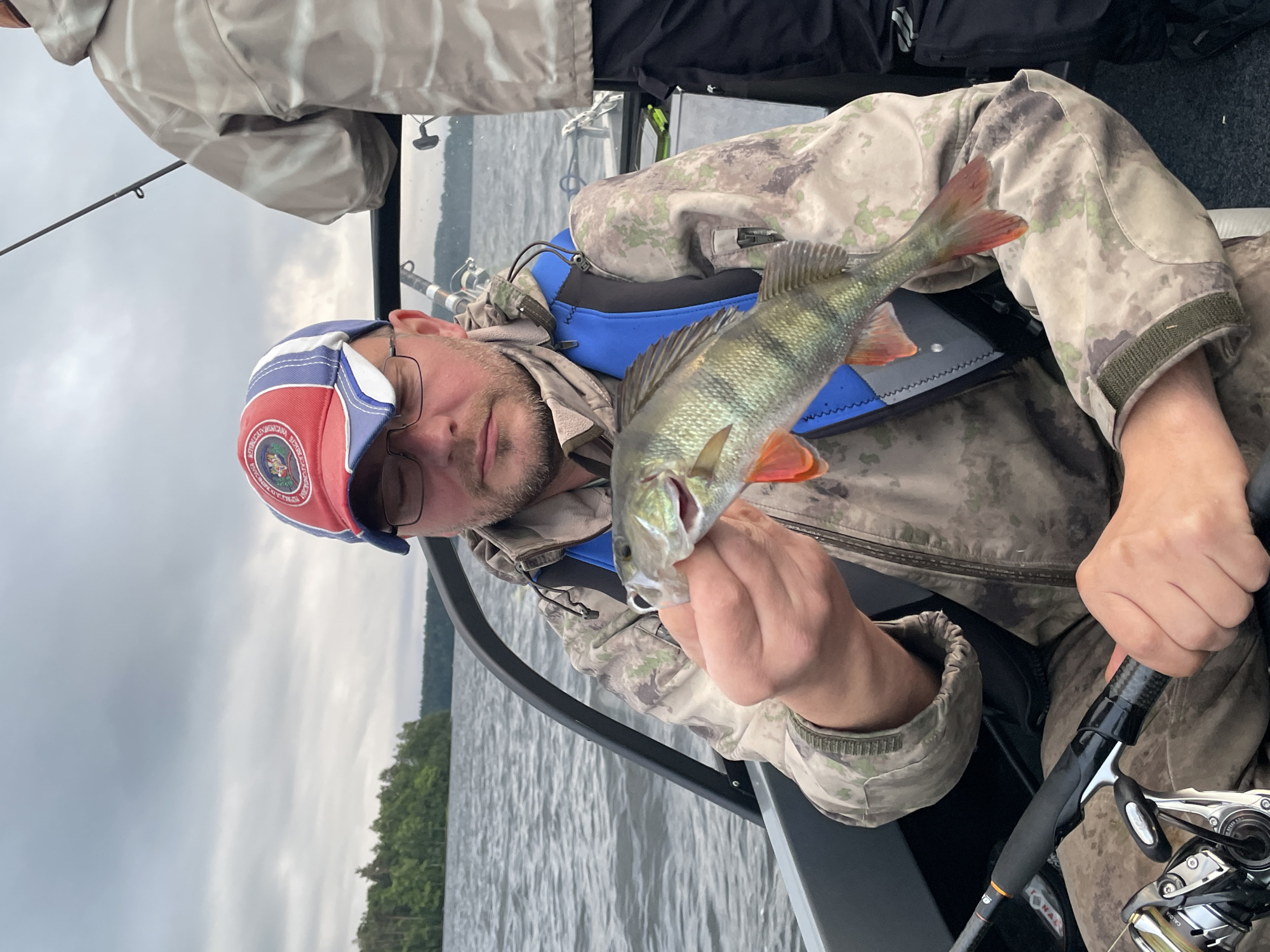 A man in a camouflage jacket and a red, white, and blue baseball cap is holding a small fish while sitting in a boat. He is wearing glasses and has a beard. Another person in a camouflage jacket is standing behind him. The boat is on a body of water, with a treed shoreline in the background. The sky is cloudy. The image was taken in the city of Yablonovka, Russia.