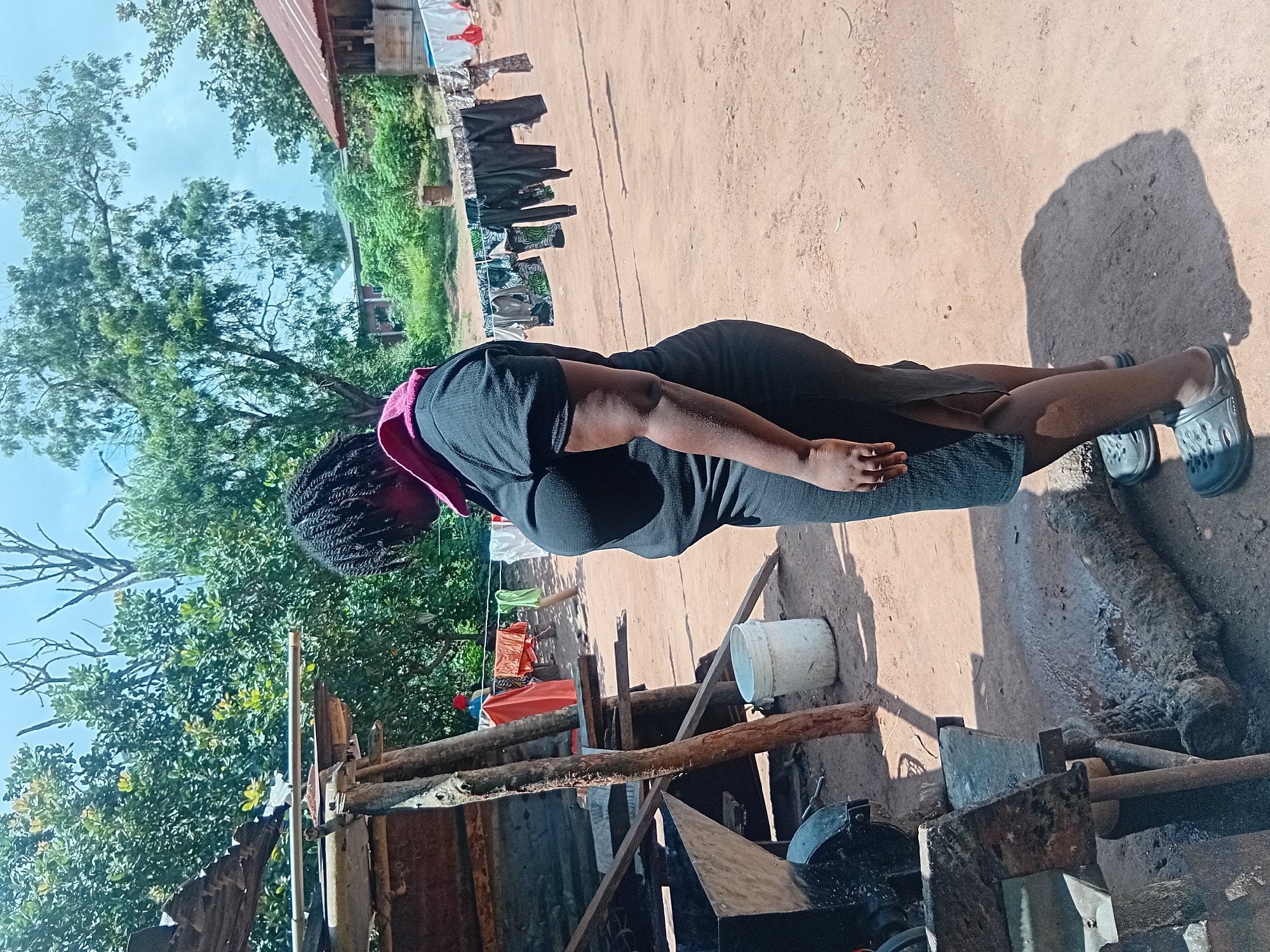 In the city of Ejule, Nigeria, a woman with dark, braided hair, wearing a black dress and dark clogs, stands facing right in a dusty, outdoor area. She appears to be looking down at a piece of machinery. Behind her, laundry is hanging on a clothesline stretched between two poles. To her left, a rustic shelter with a corrugated metal roof is visible, and further back, more buildings and lush green trees form the background. The ground is predominantly dry, sandy soil, with some scattered debris and shadows cast by the sunlight.