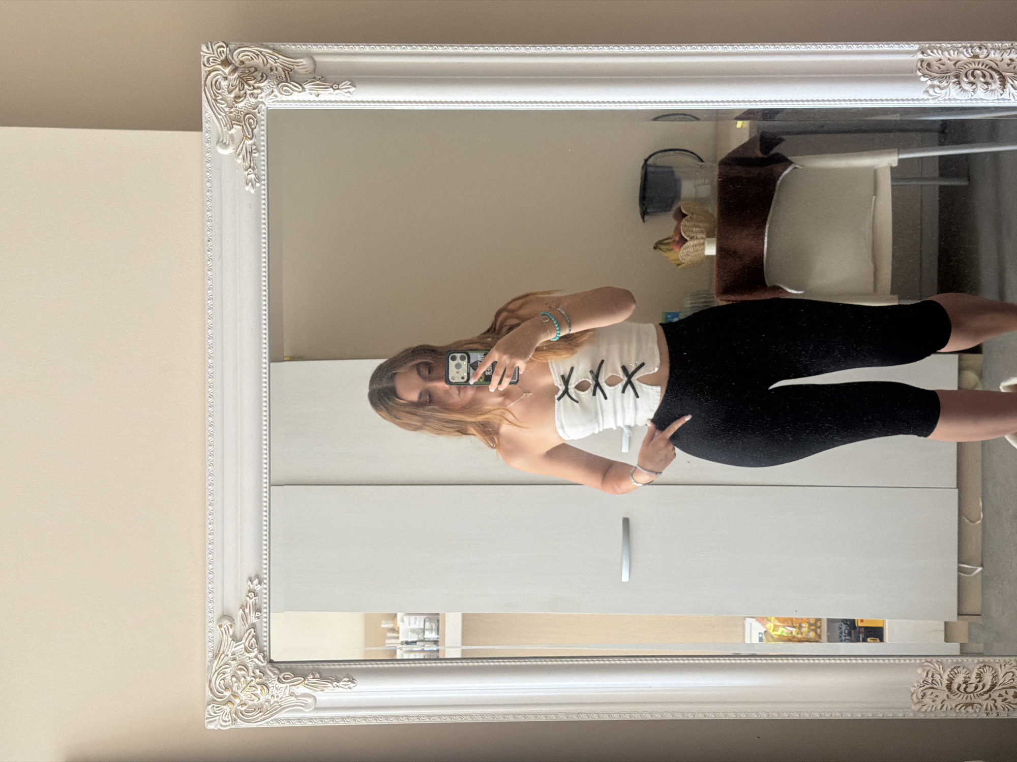 A young woman with long, wavy brown hair is taking a mirror selfie in what appears to be her bedroom in Sofia, Bulgaria. She is wearing a white strapless top with black bow details and black cropped leggings. She is standing in front of a large, ornate white mirror, which reflects her and the room behind her. The room features a white wardrobe to her right, and shelves filled with various products to her left. In the reflection, a small table with a fruit basket and a kettle is visible.