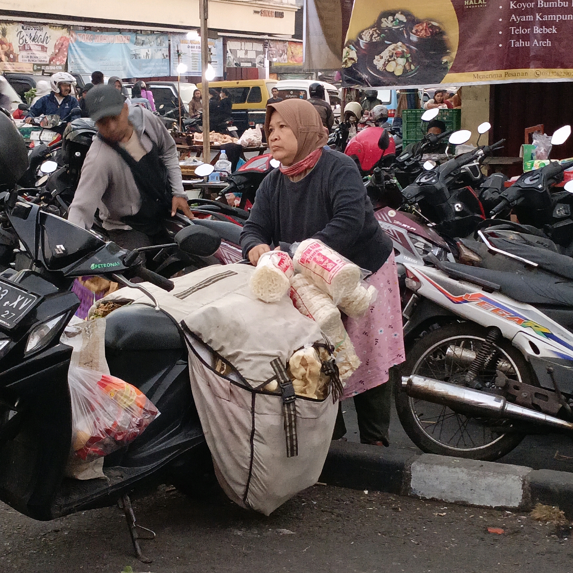 The image depicts a street scene in Salatiga, Indonesia, likely a market or a roadside stall area given the presence of parked motorcycles and vendors. In the foreground, a woman wearing a brown hijab and a pink floral skirt is seated on a motorcycle. She is holding several plastic-wrapped bundles of what appear to be snacks or food items. A large canvas bag, also on the motorcycle, contains more goods. Other motorcycles are parked closely around her.
In the background, various vehicles, including cars and more motorcycles, are visible. Several people are present, some interacting with stalls or riding motorcycles. Banners with Indonesian text and food imagery suggest commercial activity. The environment appears to be an urban setting with a paved road and a curb. The overall impression is one of a busy, local marketplace where people are engaged in shopping and transportation.