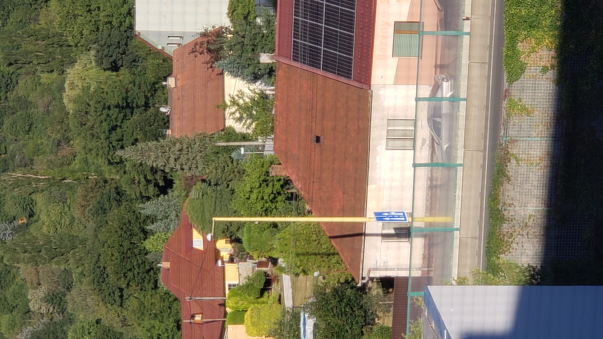 The image depicts a street view in Prešov, Slovakia, characterized by residential buildings with red tiled roofs, lush green trees, and a section of a road with a barrier. A silver car is visible on the road, partially obscured by a transparent barrier that separates the road from the foreground. A tall yellow lamppost stands near the barrier, with a blue and white directional sign at its base. The buildings in the background are situated on a slope, with more dense greenery covering the upper parts of the hills. One building has solar panels on its roof. The scene suggests a suburban or semi-urban environment, likely during daylight hours given the natural lighting.